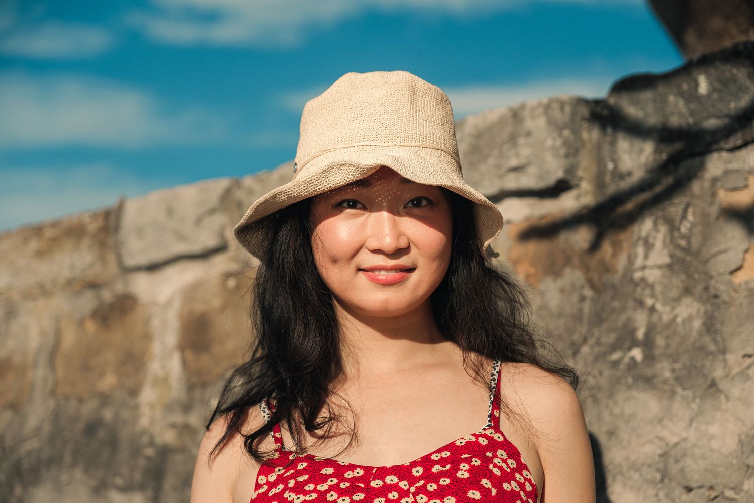 Young Asian woman in a beige bucket hat and red floral dress smiling outdoors under blue sky.