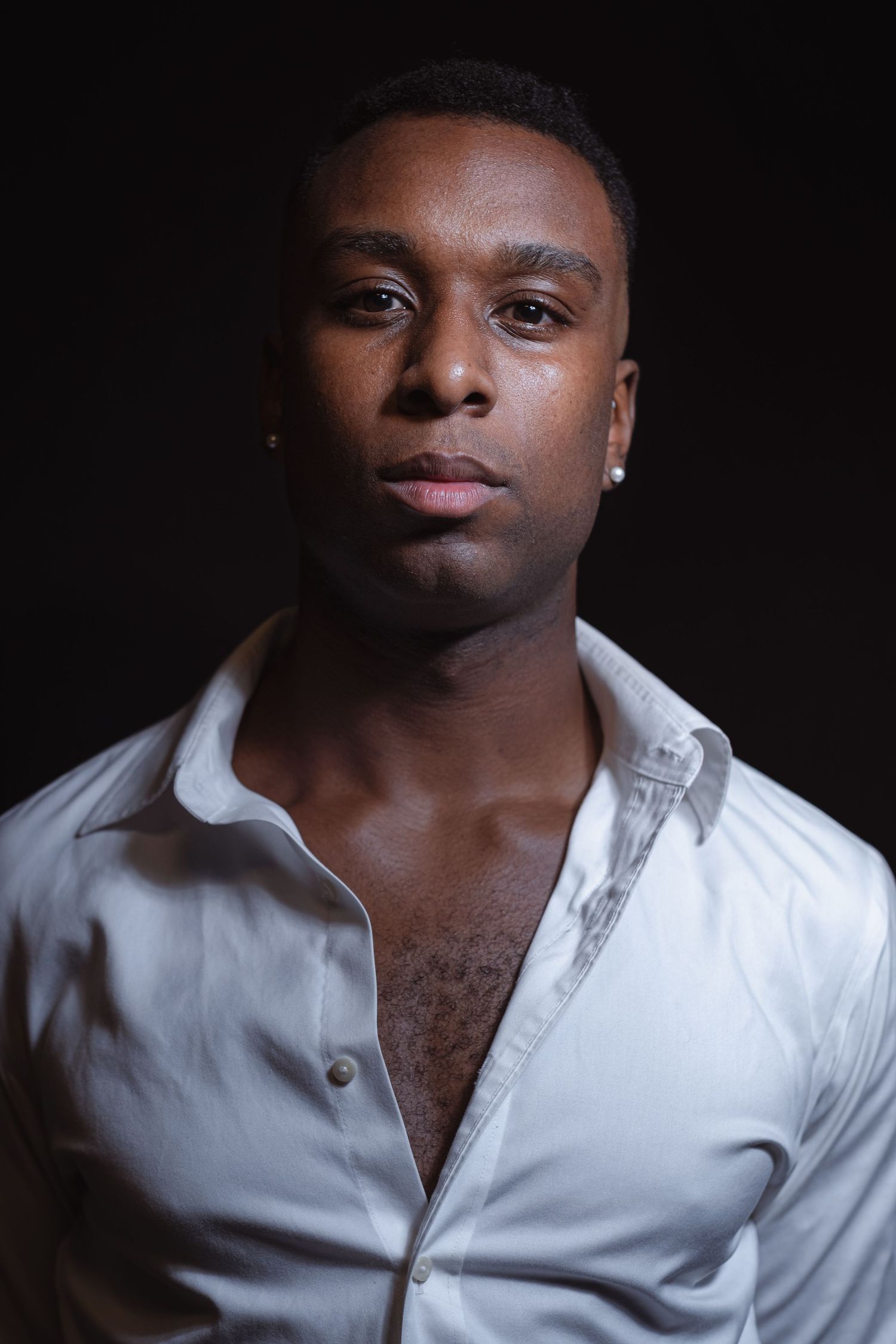 A young Black man in an open white shirt poses against a dark background in a dramatic studio portrait.