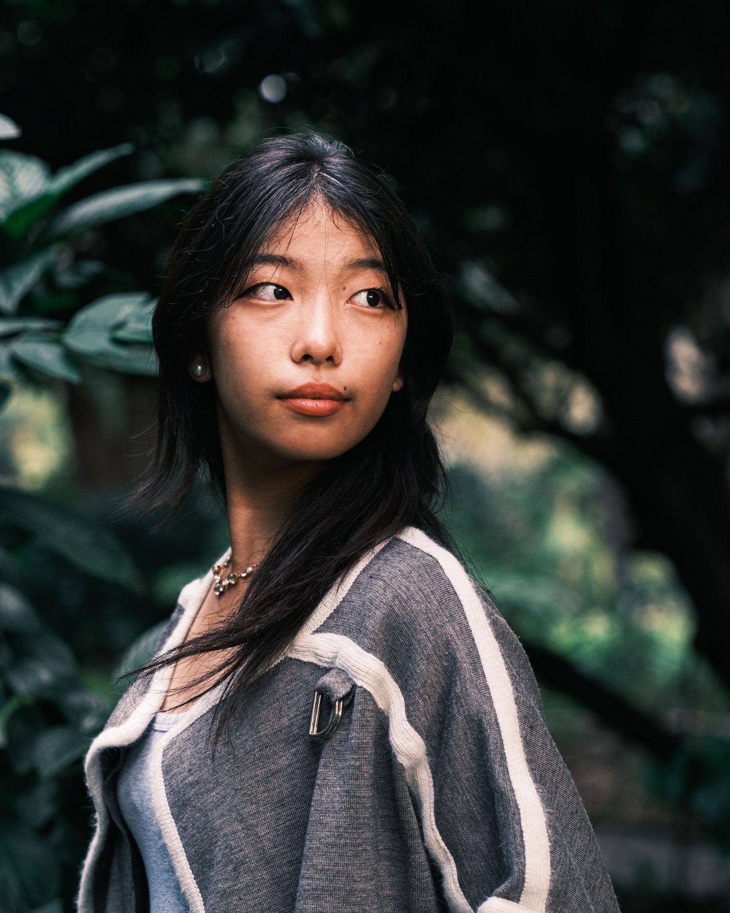 A young Asian woman in a gray cardigan poses outdoors with lush green foliage in the background.