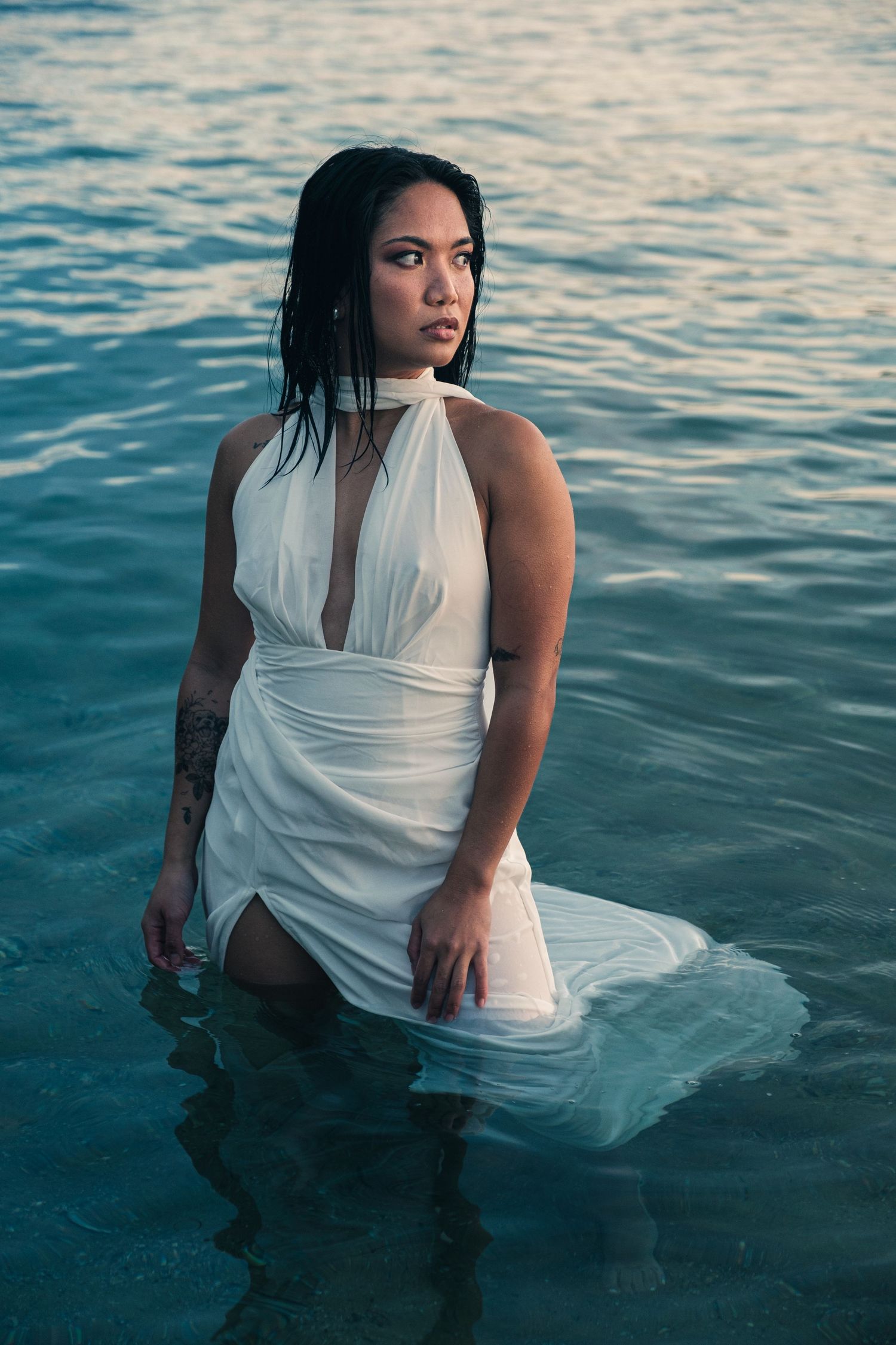 A woman in a wet white halter dress stands in ocean water at dusk in an artistic fashion photo.