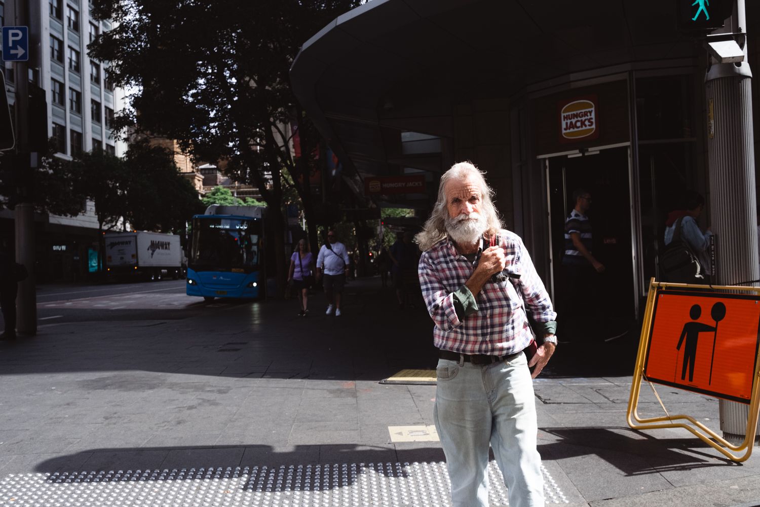 An elderly man with a long white beard stands on a sunny urban sidewalk near a construction sign.