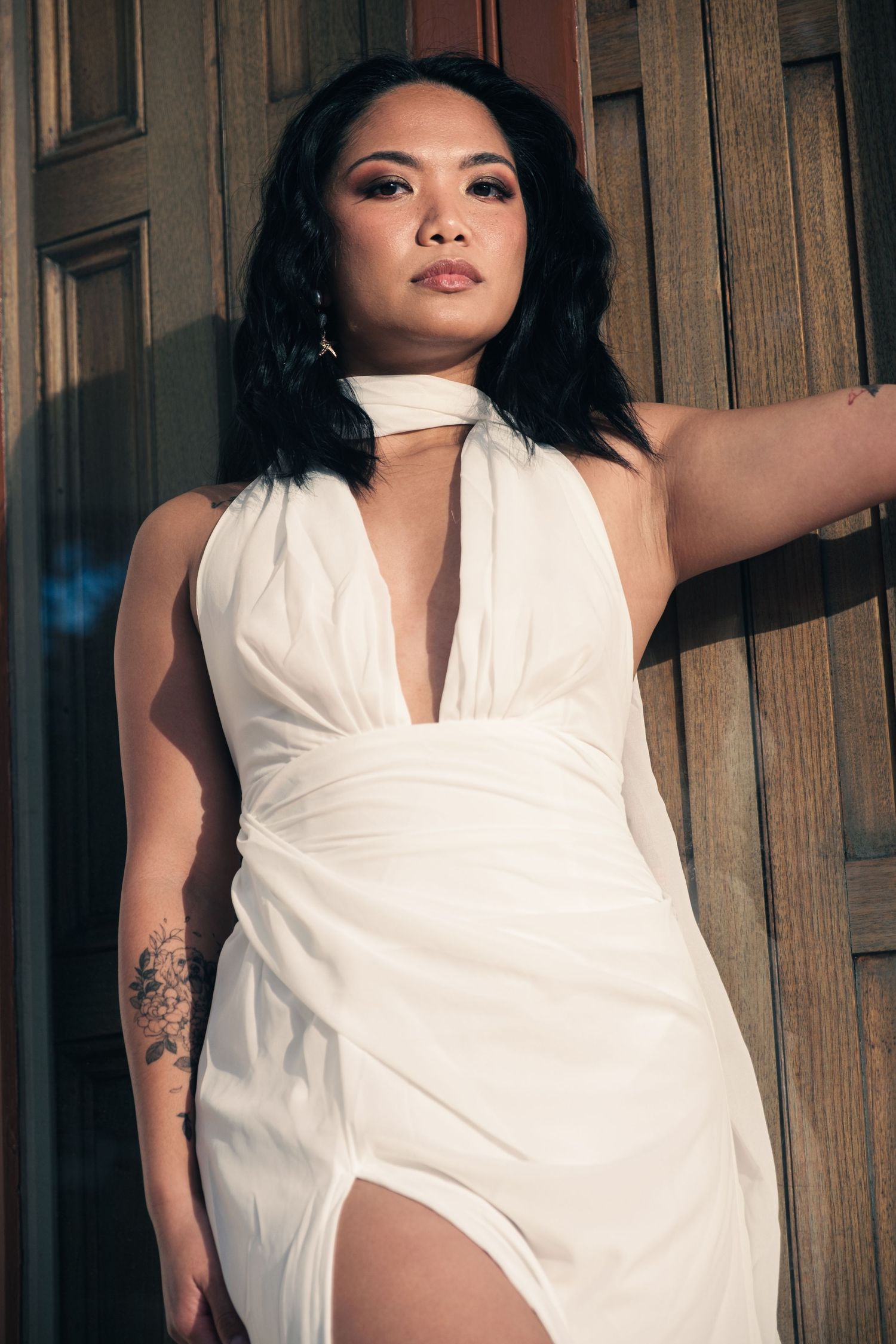 A woman in a white halter dress with deep V-neckline poses against a wooden door in moody lighting.