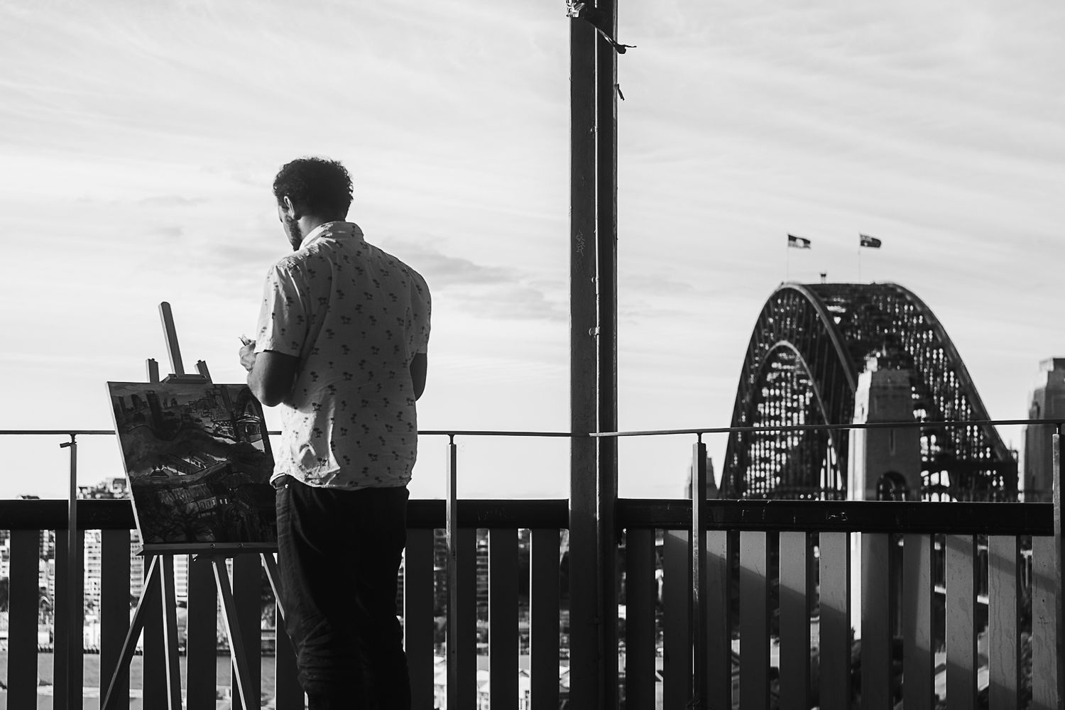 Artist painting on easel with Sydney Harbour Bridge visible in black and white background.