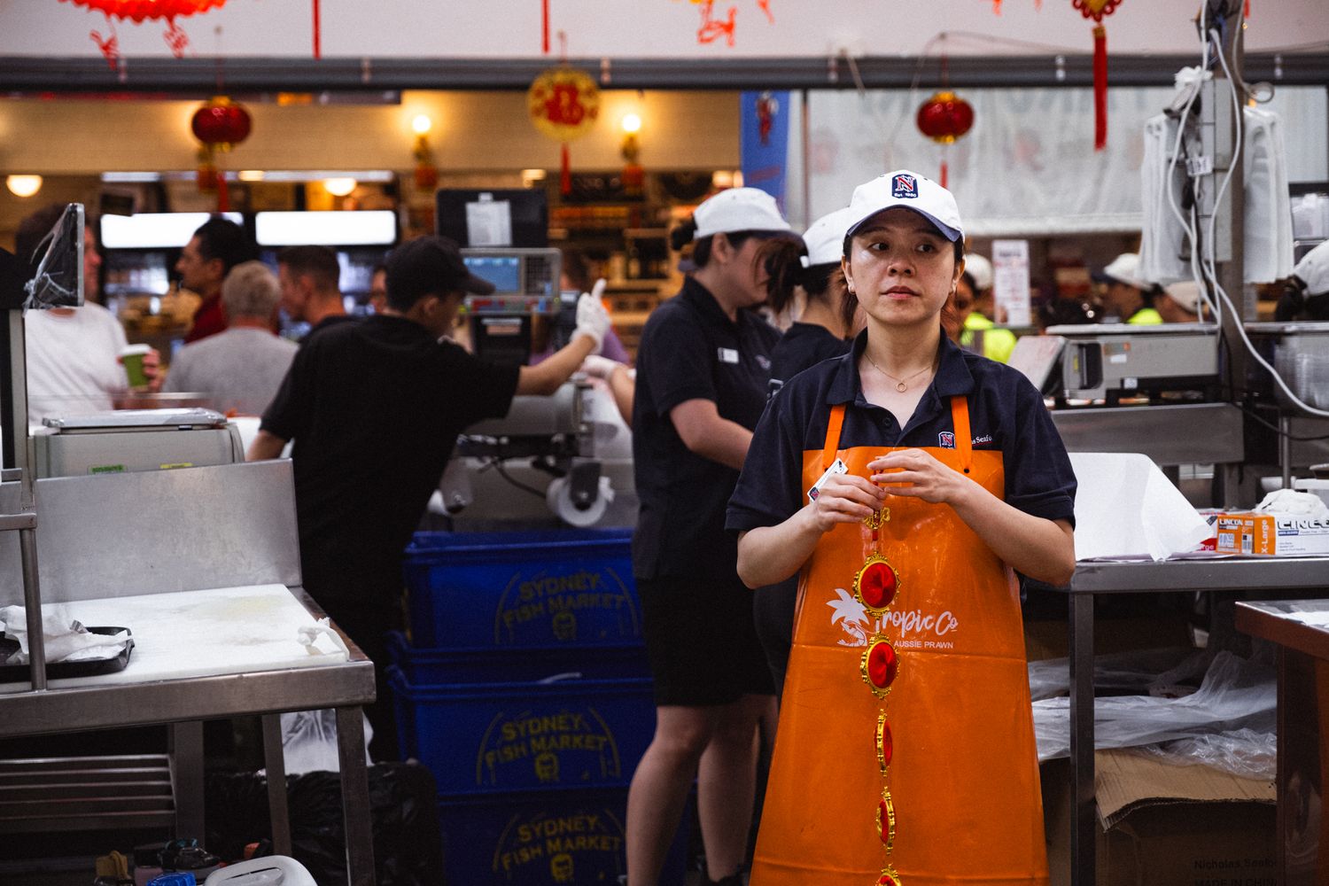 A young female worker in an orange apron stands in a busy seafood market decorated with red lanterns.