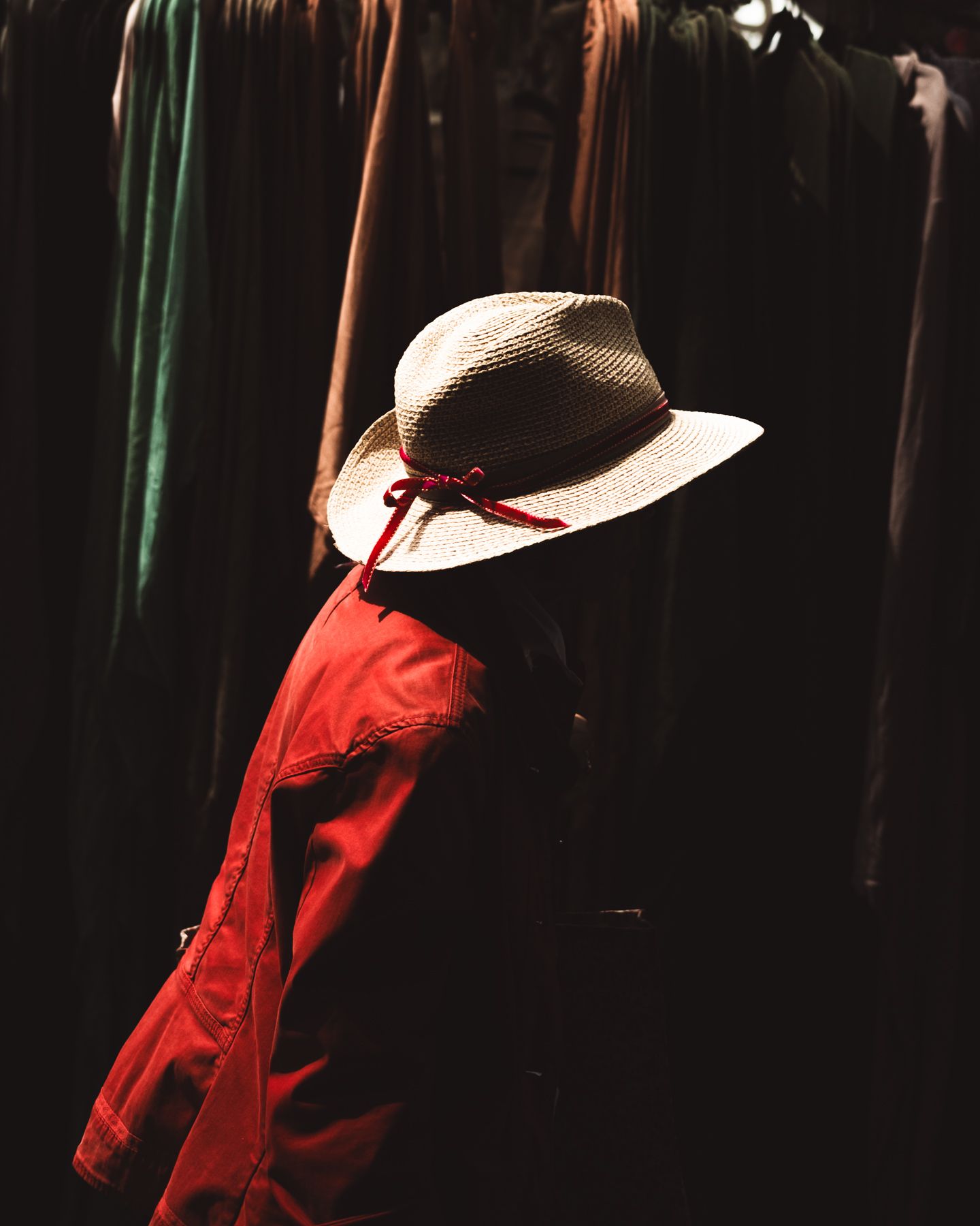 A person in a red jacket and straw hat with red ribbon browses through a rack of dark clothing.