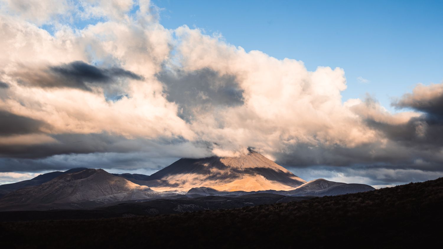 Dramatic clouds billow over a volcanic mountain peak at sunset with warm golden light illuminating the landscape.