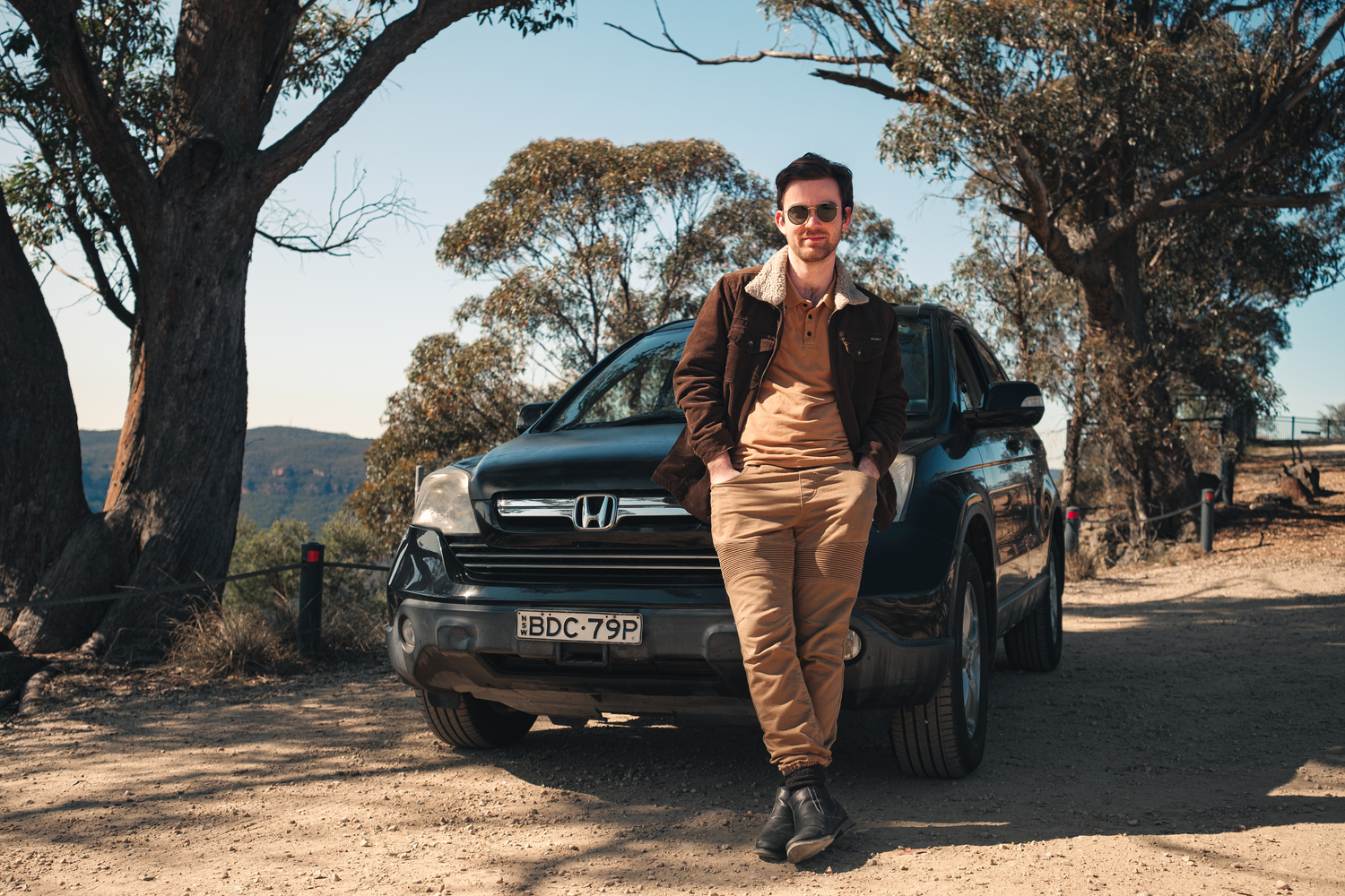 Young man in brown jacket and tan pants leaning against a black Honda CR-V on a dirt road surrounded by trees.