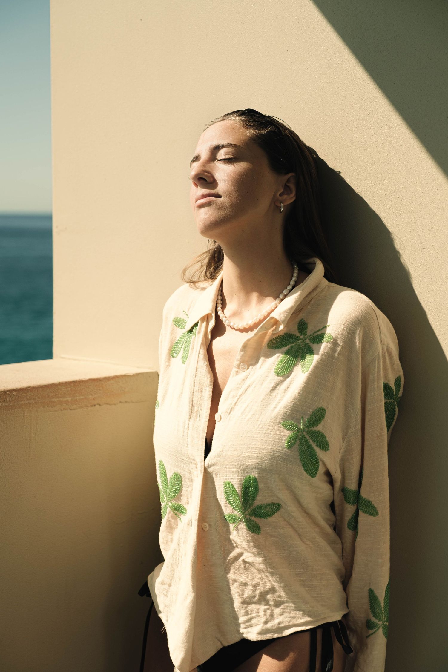 Woman in floral shirt leaning against wall with eyes closed near the ocean.