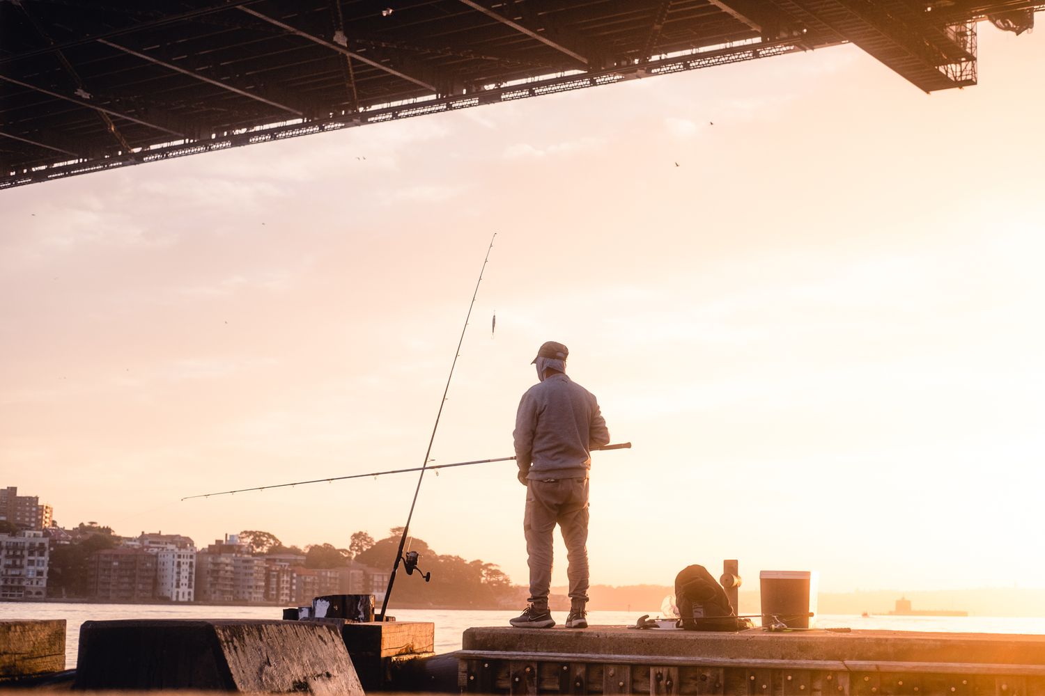 A lone fisherman silhouetted against a golden sunset sky casts his line from a waterfront pier.