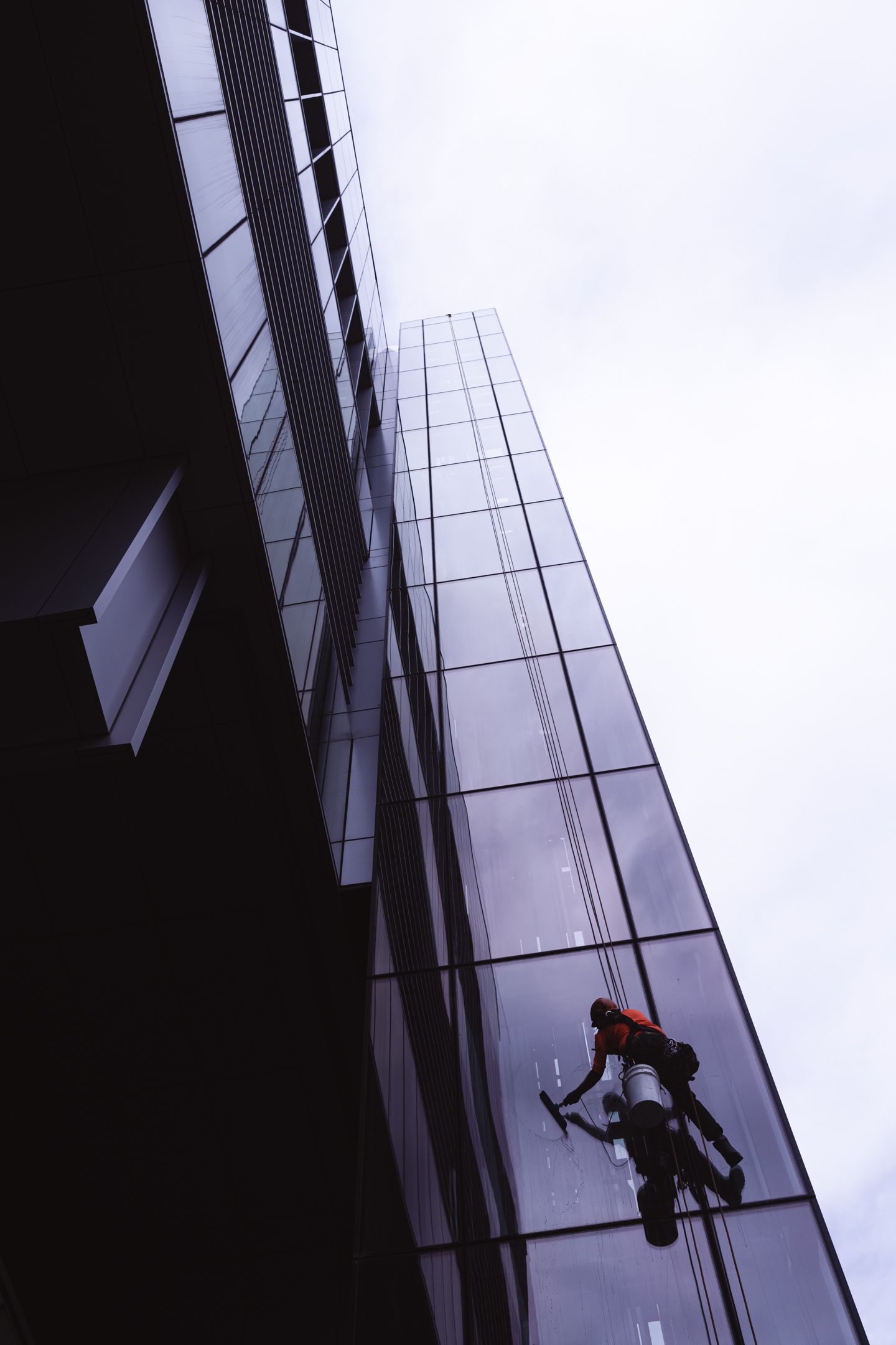 Window cleaner rappelling down a tall glass skyscraper facade against an overcast sky.