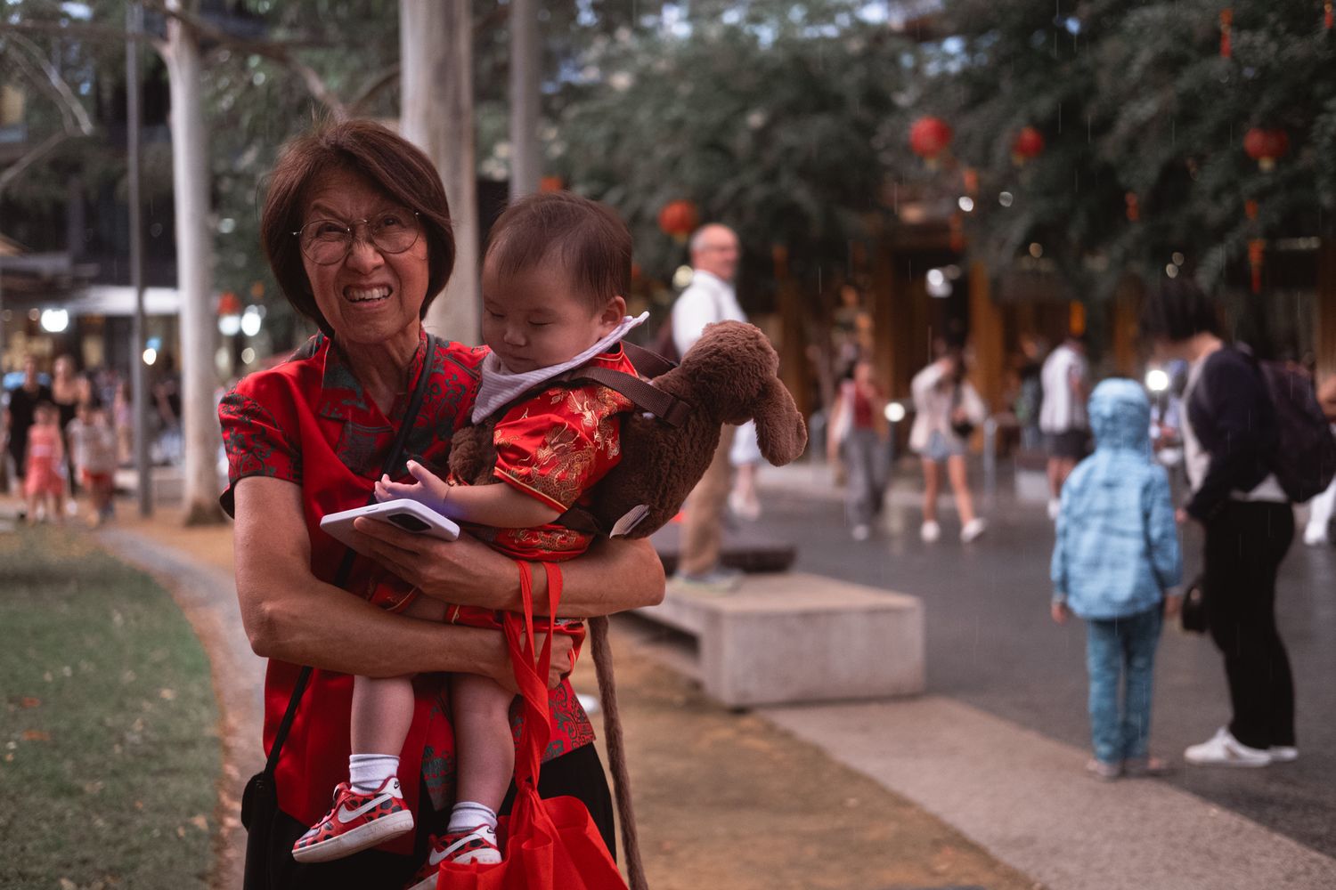 Smiling woman in red Chinese dress holding baby outdoors at festive evening event.