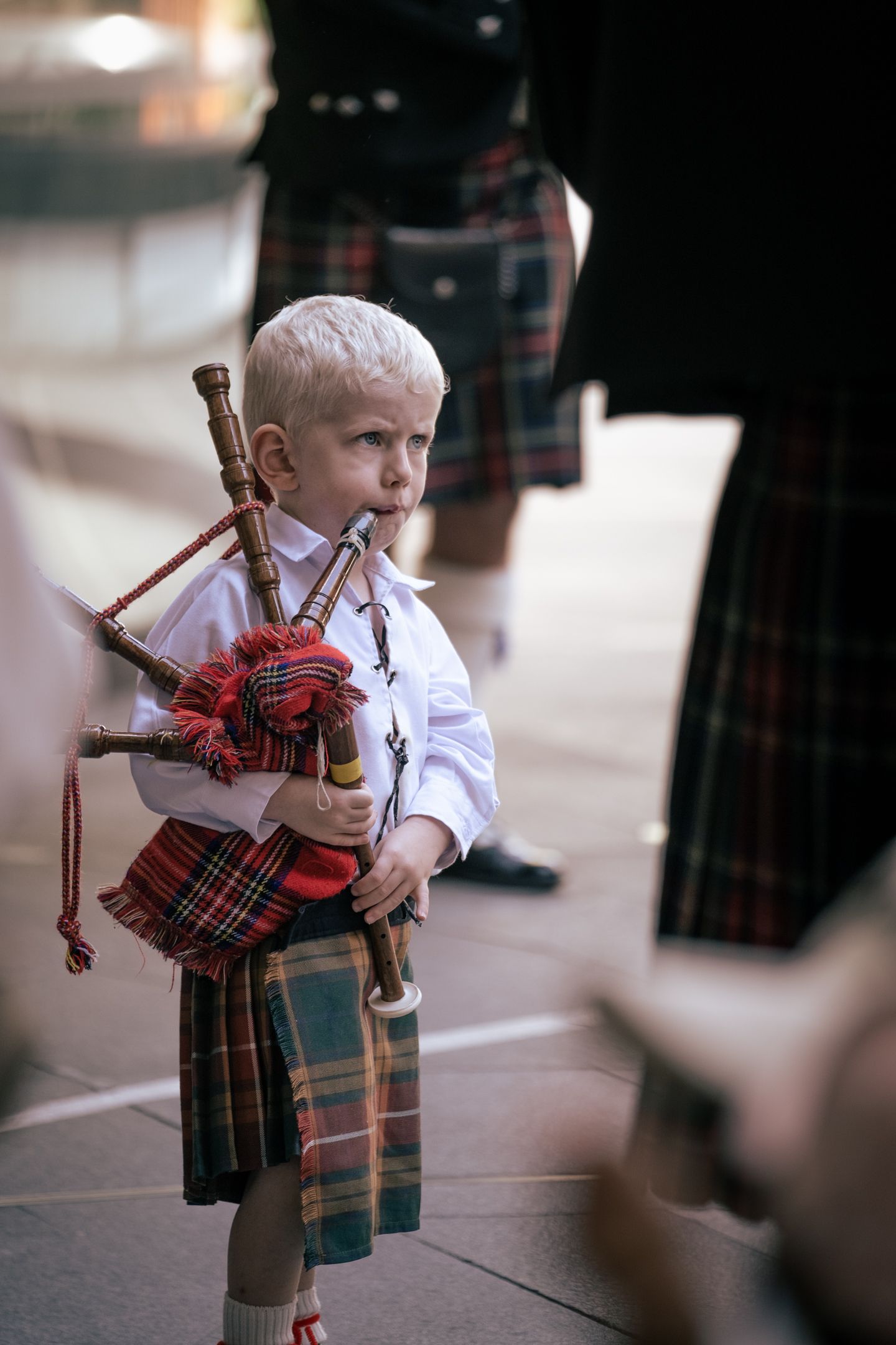 Young blonde boy in tartan kilt playing bagpipes at a traditional Scottish parade.