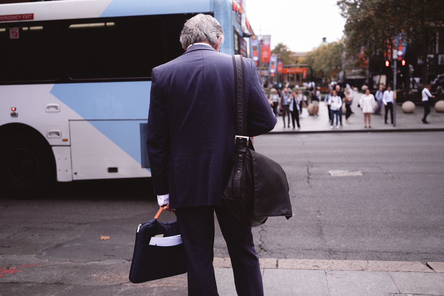 Elderly businessman in blue suit carrying bags, standing on city street with double-decker bus nearby.
