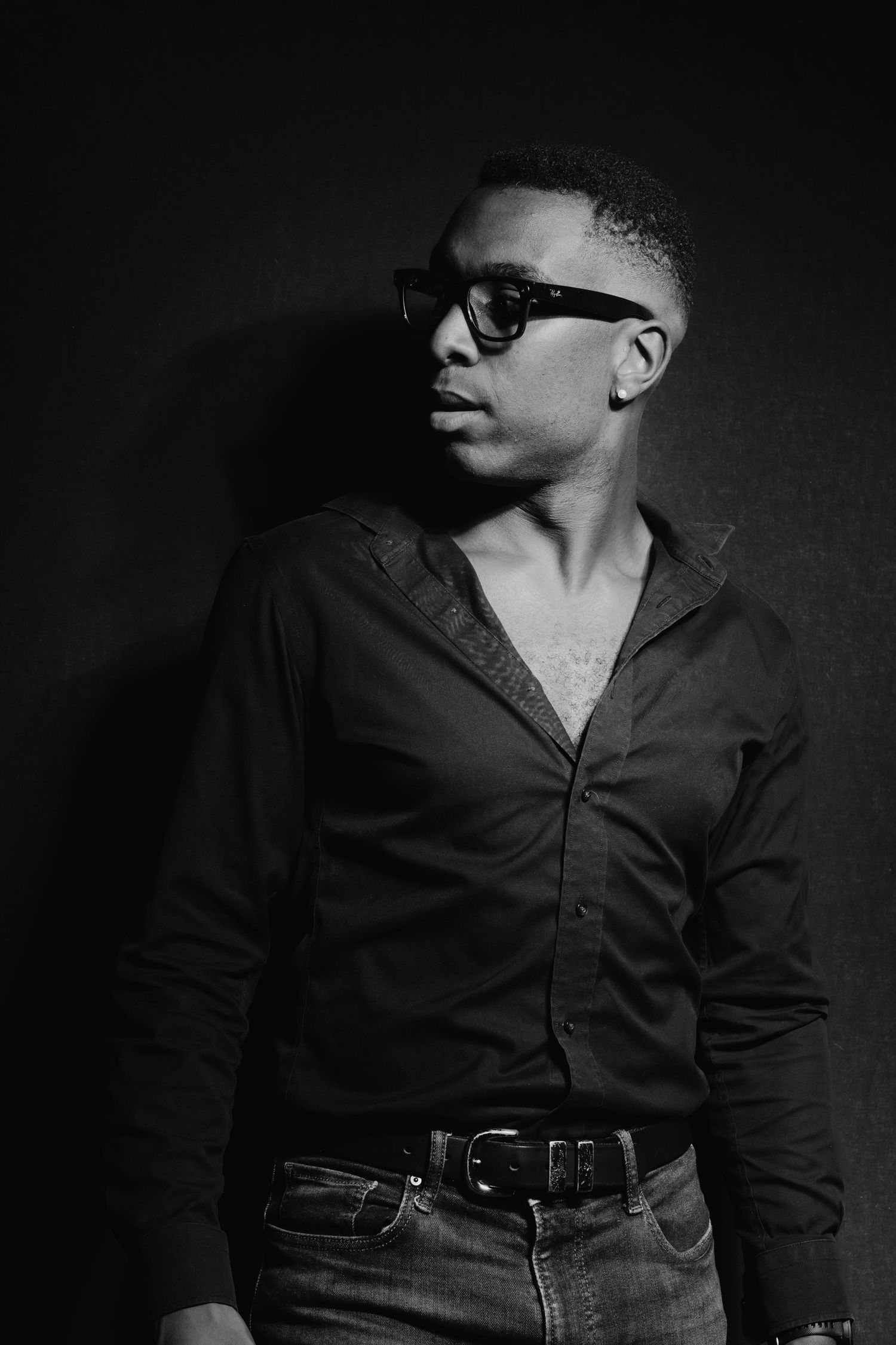 Black and white portrait of a young man in sunglasses and open black shirt against dark wall.