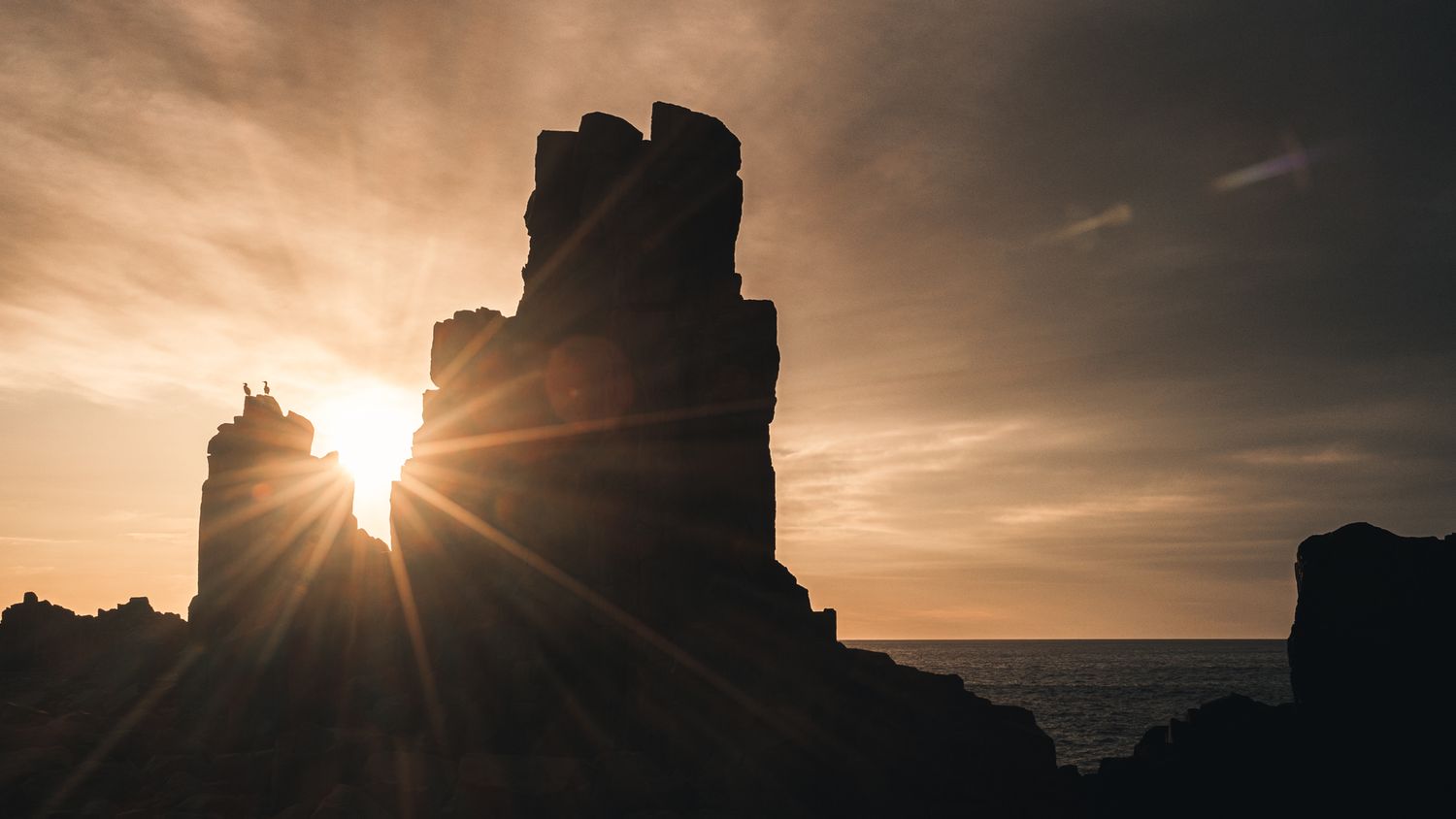 Silhouetted rock formation with sun rays bursting through at sunset over the ocean.