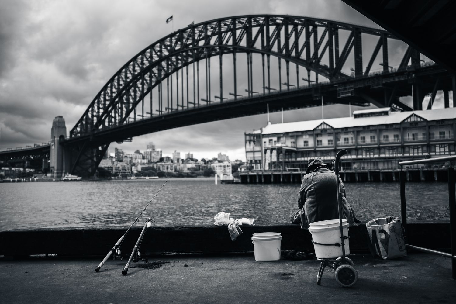 Person fishing alone near Sydney Harbour Bridge on a cloudy day in black and white.