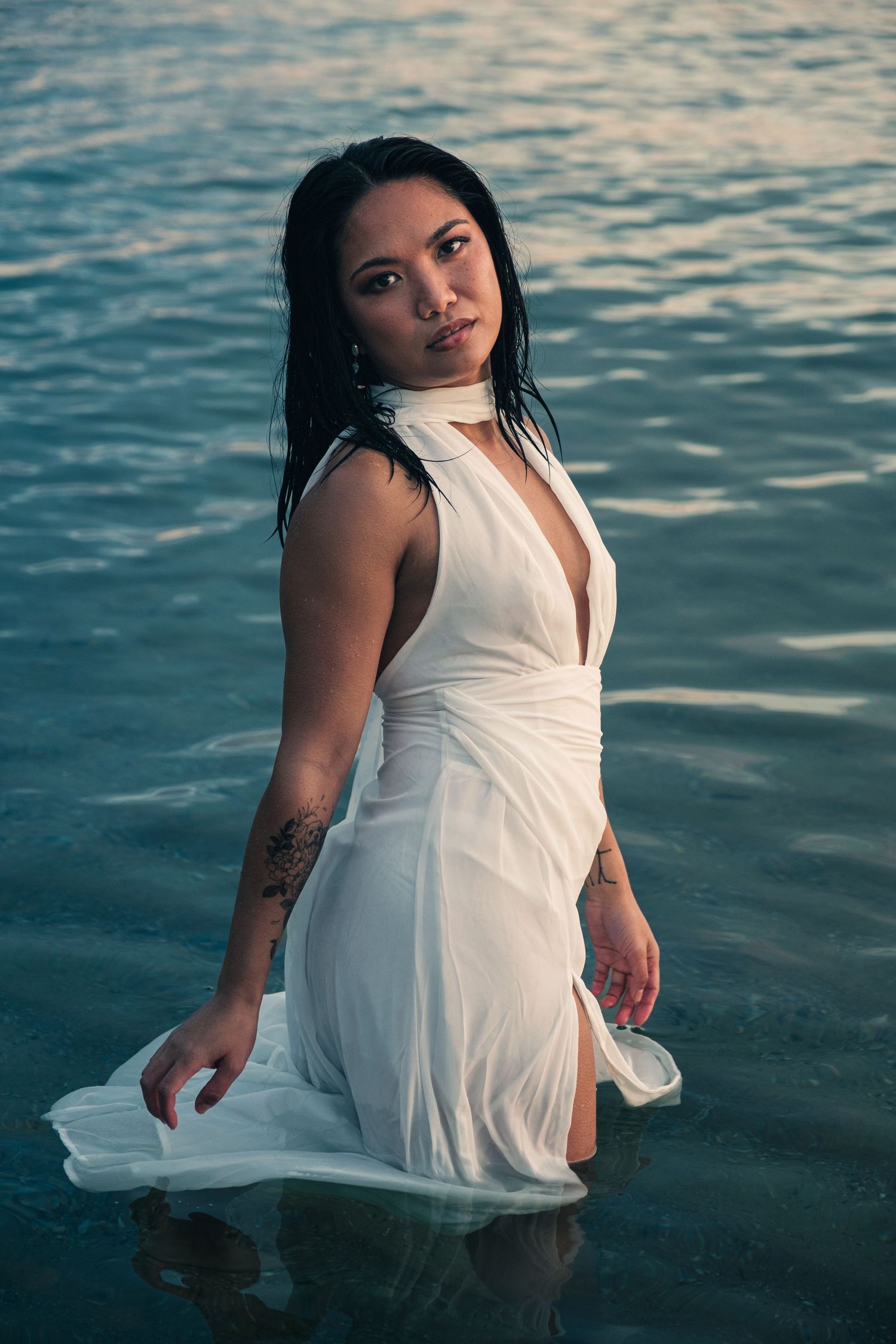 A woman in a wet white halter dress stands in shallow ocean water at dusk.