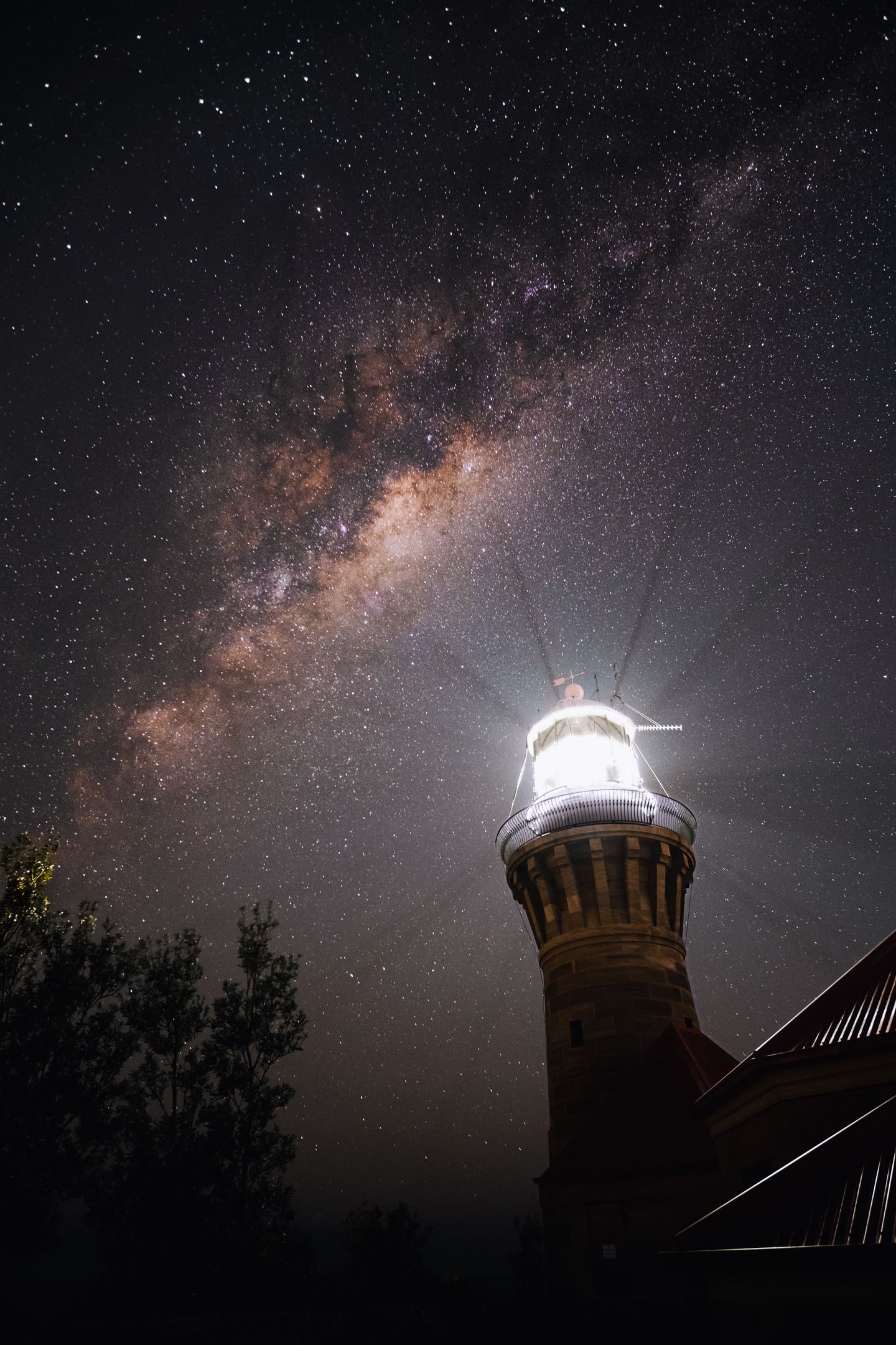 Lighthouse glowing beneath a stunning Milky Way galaxy in a starry night sky.