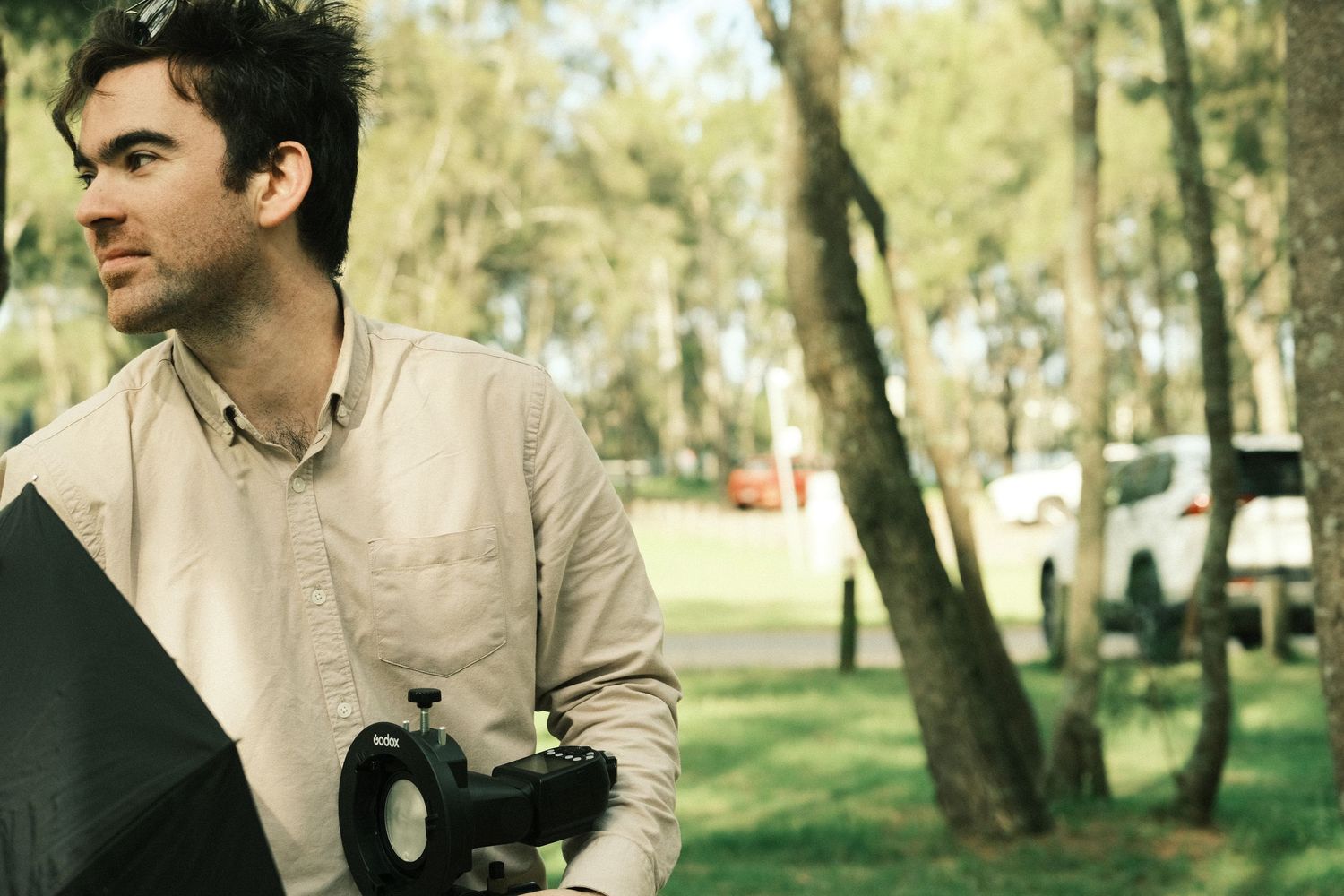 Man holding camera equipment looking away in sunlit park with trees in background.