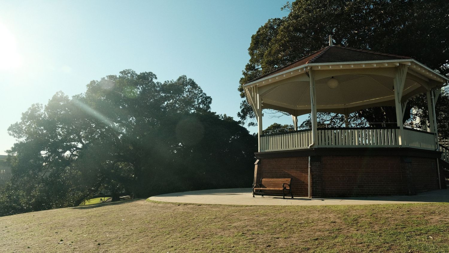 Sunlit Victorian gazebo bandstand on a hill surrounded by lush green trees in a peaceful park setting.