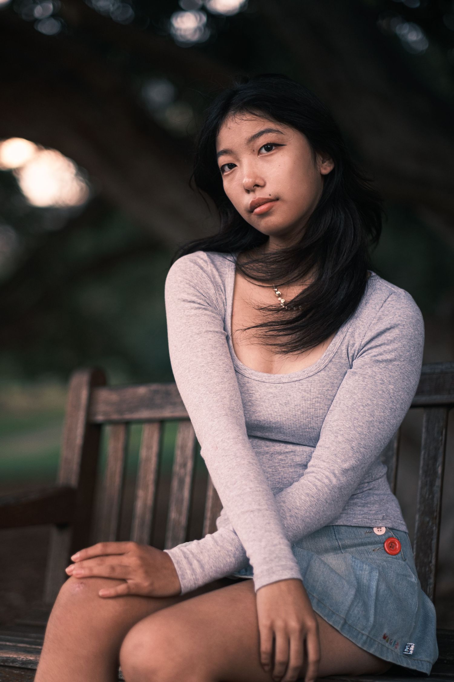 A young Asian woman in a gray long-sleeve top and denim skirt sits on a wooden bench outdoors.