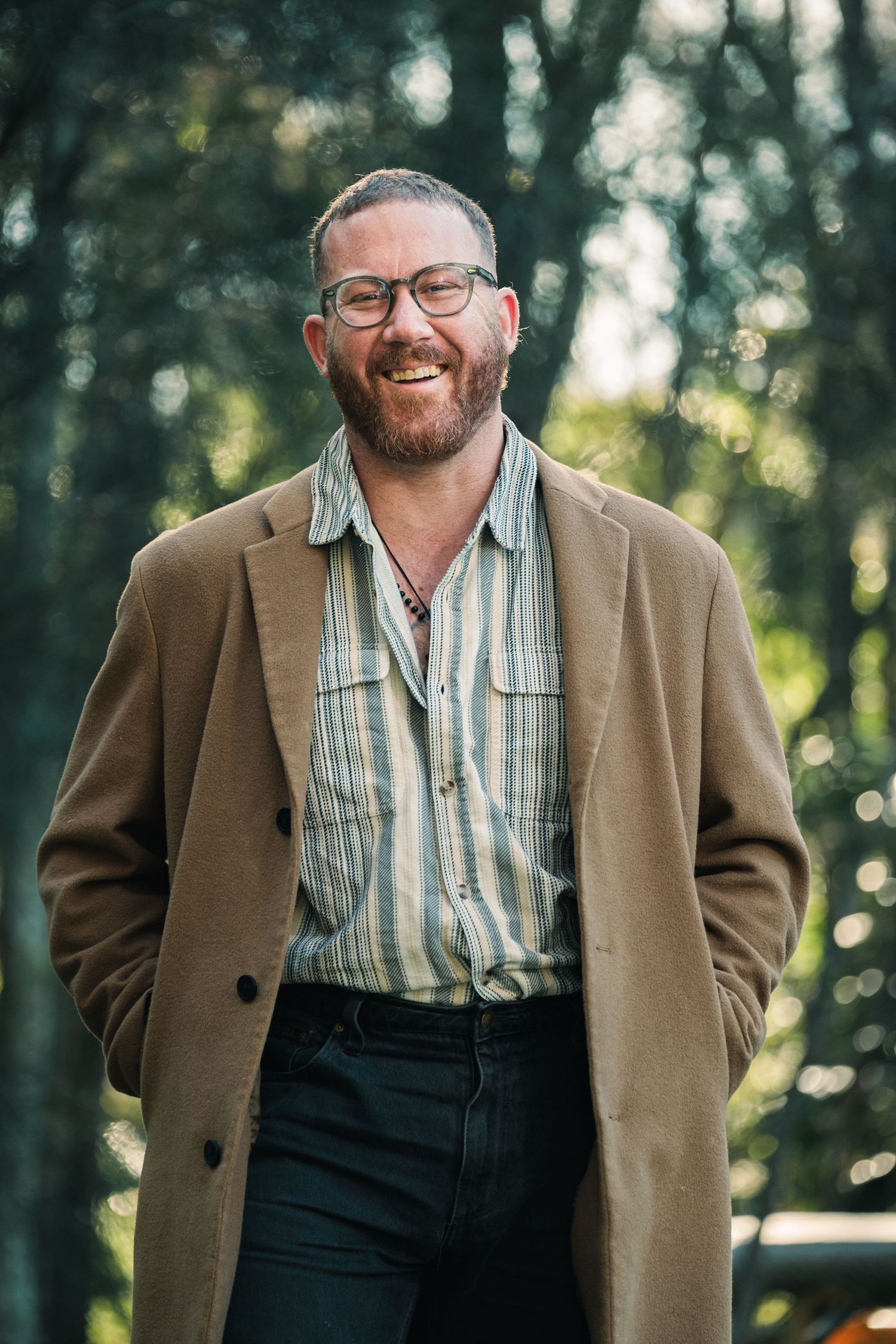 Smiling bearded man wearing glasses, striped shirt, and tan coat standing outdoors among green trees.