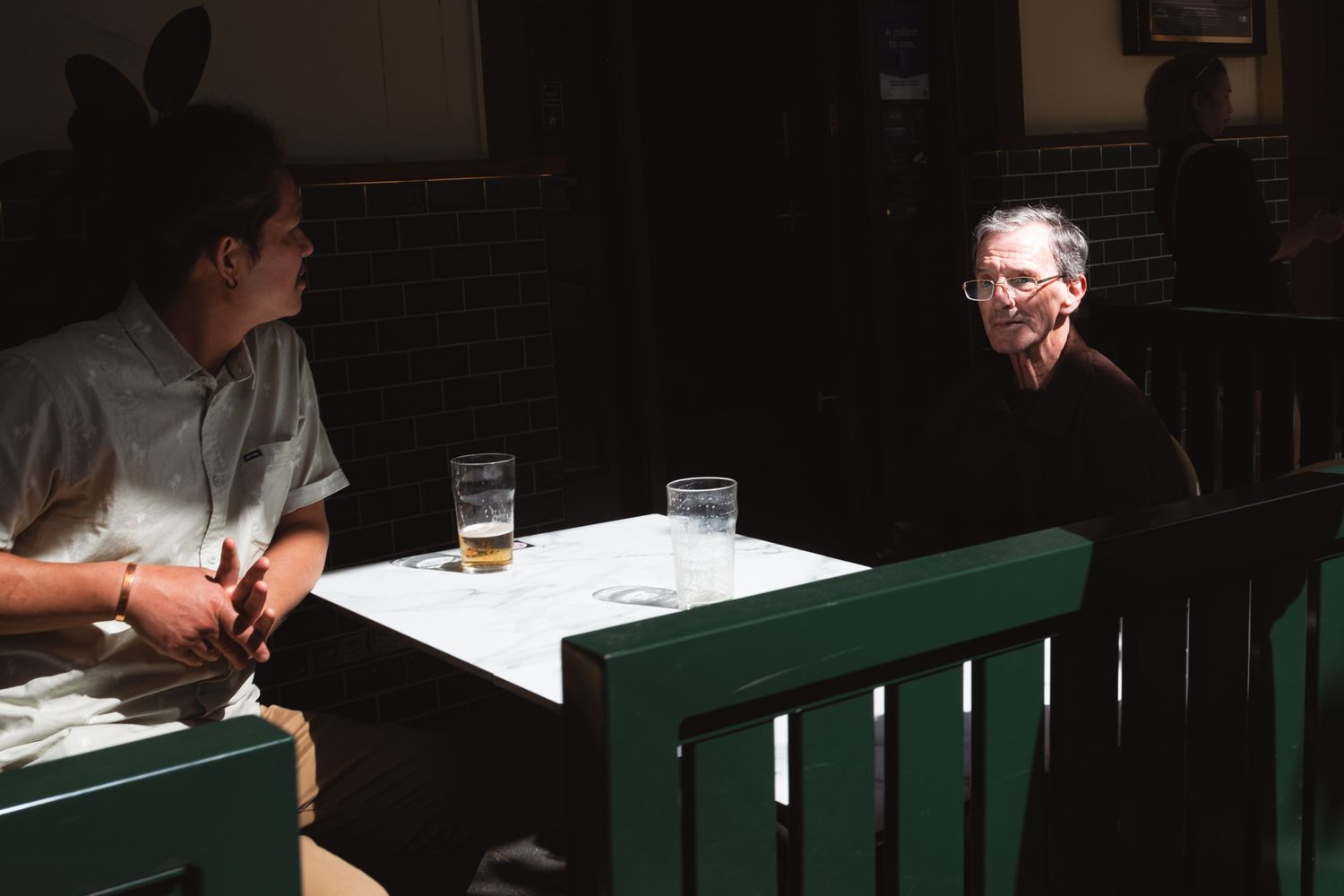 Two men sit across from each other at a green table with drinks in a dimly lit bar.