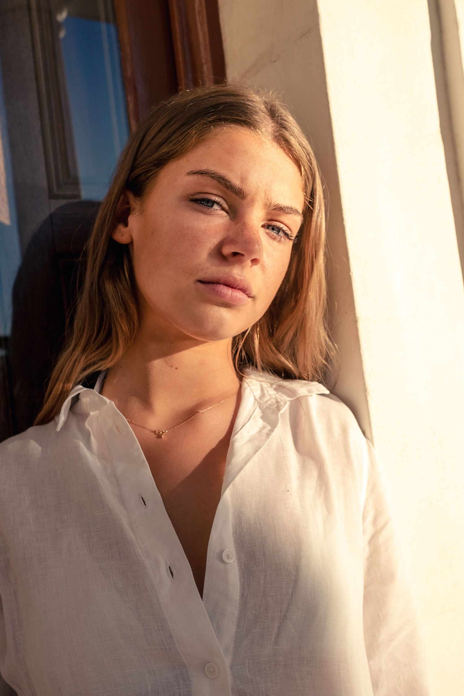 A young blonde woman in a white linen shirt bathed in warm golden sunlight near a bright window.