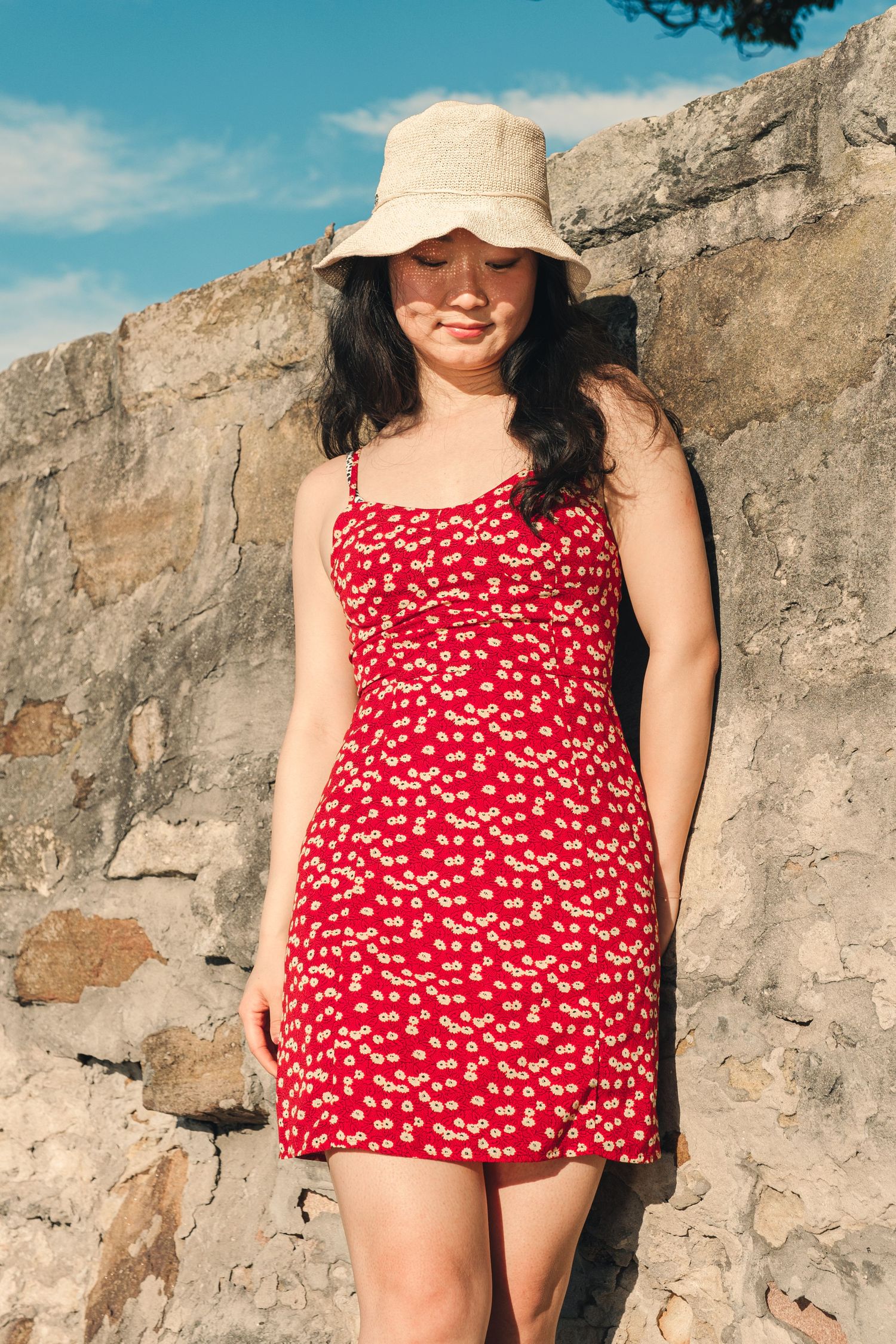 Young woman in a red floral sundress and beige bucket hat standing by a stone wall outdoors.