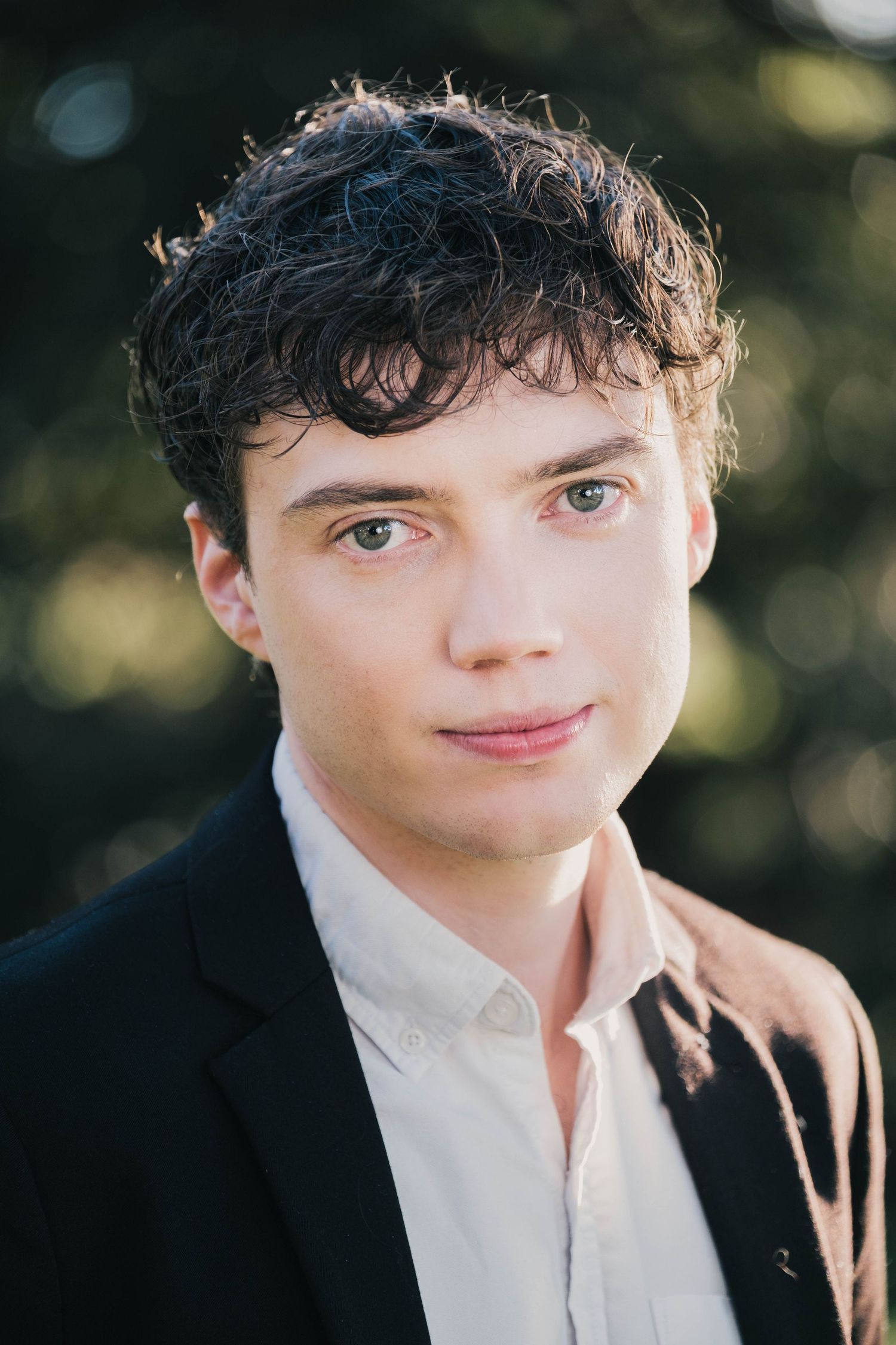Young man with curly dark hair wearing a black blazer and white shirt, posing outdoors with soft natural lighting.