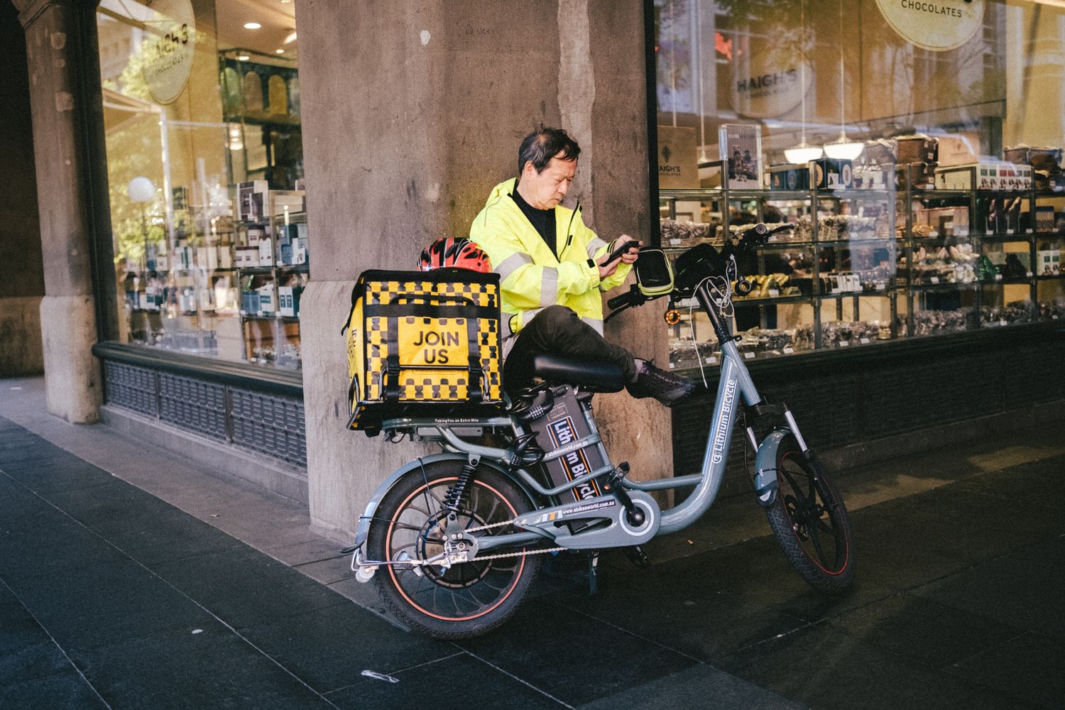 A delivery rider in a yellow jacket checks his phone while sitting on an electric bike outside a shop.