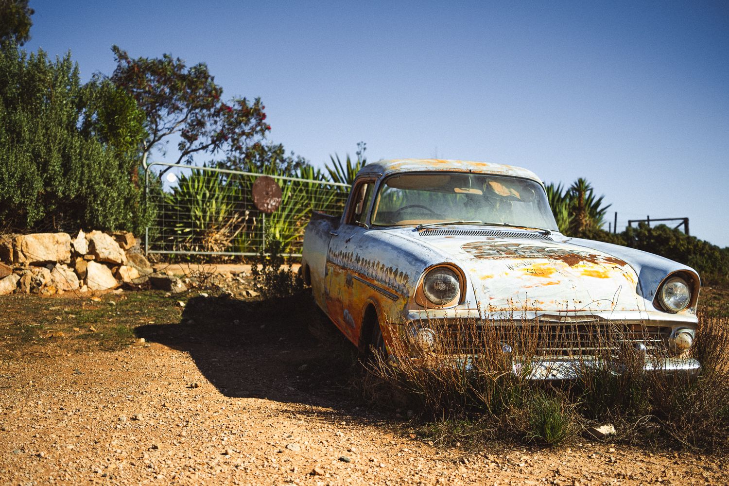 Abandoned rusty vintage American car sits weathered on a dusty dirt road under a clear blue sky.