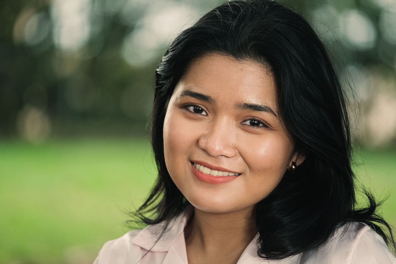 Smiling Southeast Asian woman with dark hair posing outdoors in a bright green park setting.