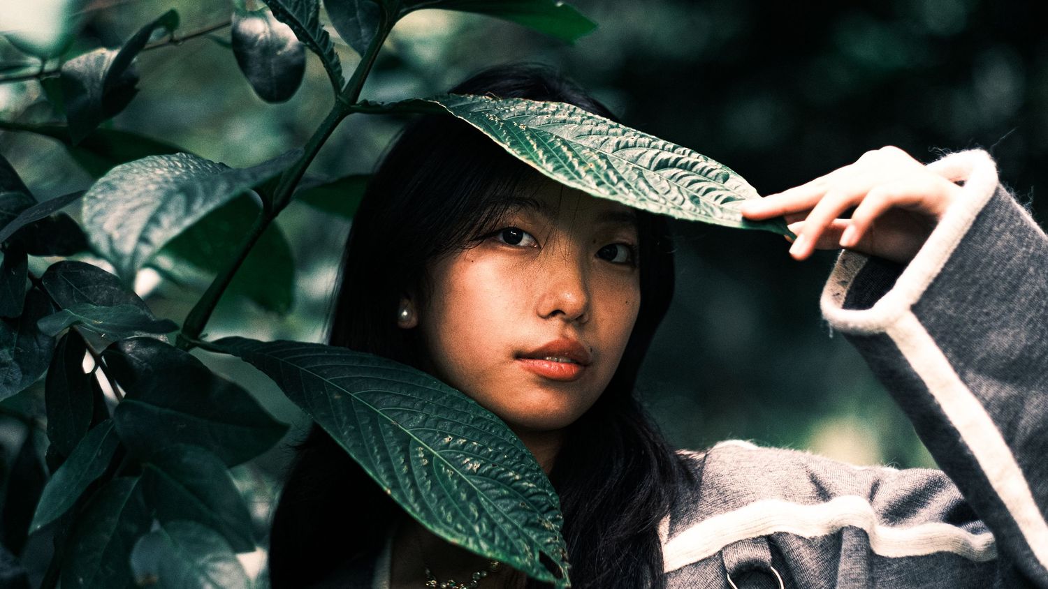 Young Asian woman with dark hair holding a large green leaf in a lush garden setting.