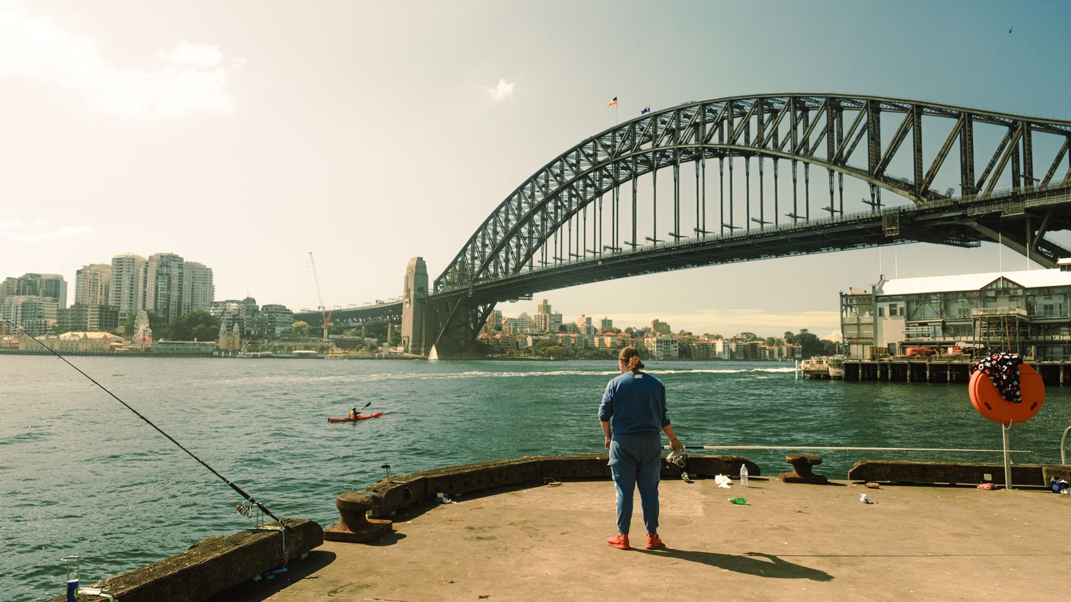 Person fishing on dock with iconic Sydney Harbour Bridge spanning the water in warm vintage tones.