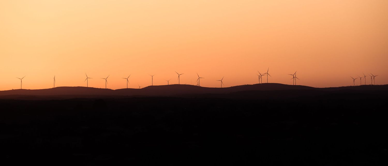Silhouetted wind turbines line rolling hills against a vivid orange sunset sky in a panoramic landscape.