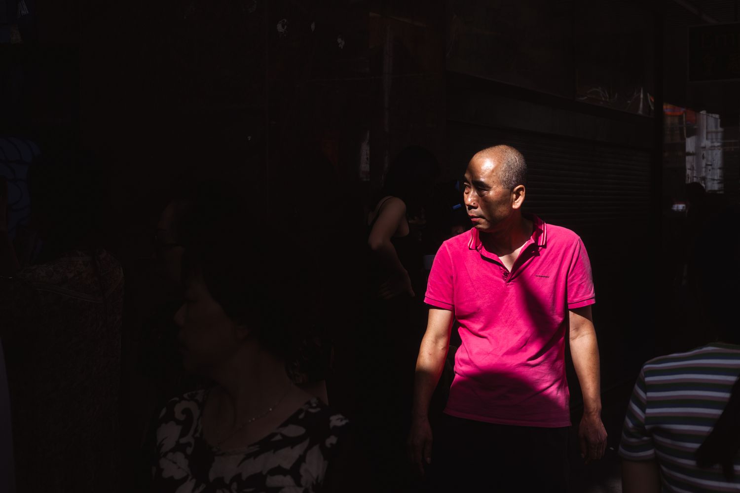 A bald man in a bright pink polo shirt stands in dramatic light against a dark background.
