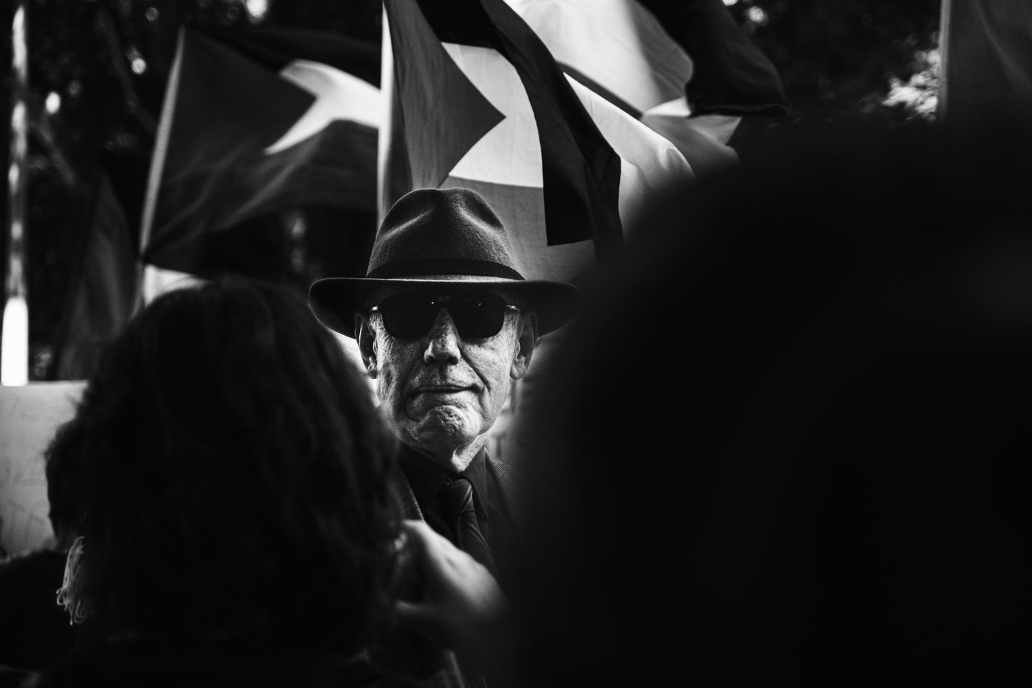 A stern elderly man in a hat and sunglasses stands among crowd members holding flags in black and white.