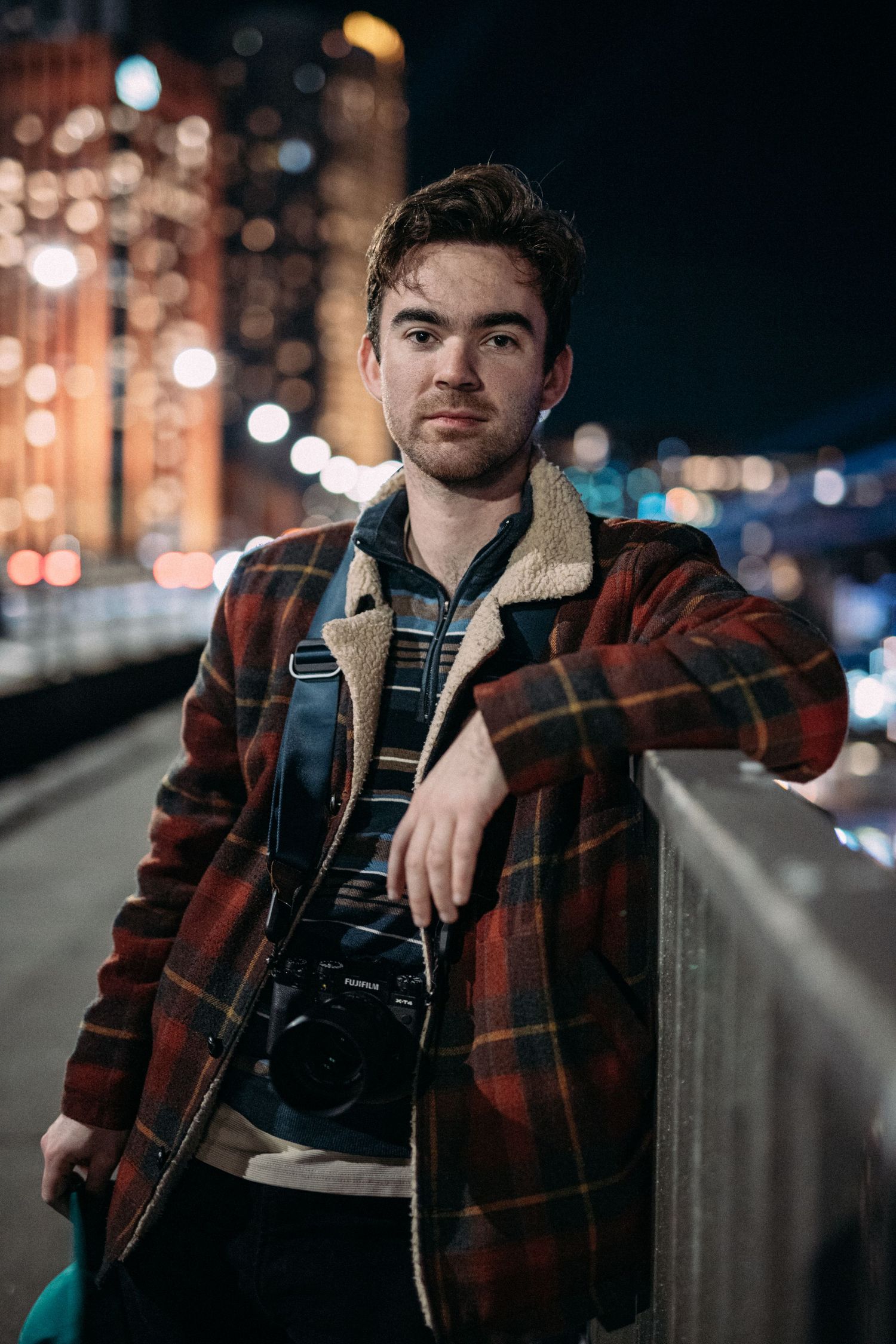 Young man in plaid jacket holding camera on city bridge at night with bokeh lights.