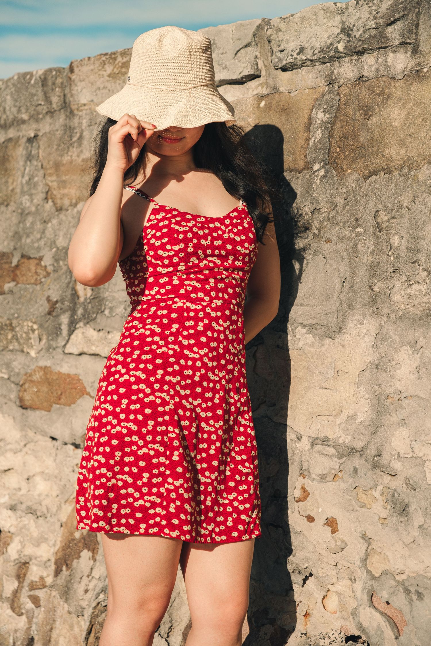 Young woman in a red floral mini dress adjusting her beige bucket hat near a rustic stone wall.
