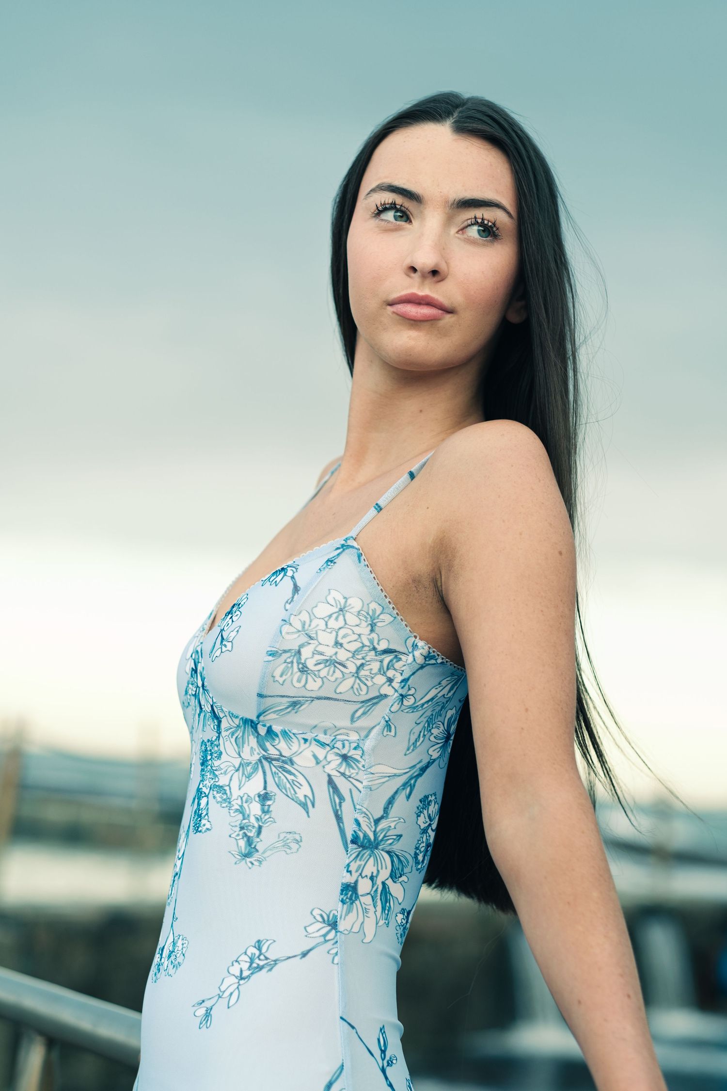 Elegant young brunette woman in a blue floral spaghetti strap dress posing outdoors at dusk.