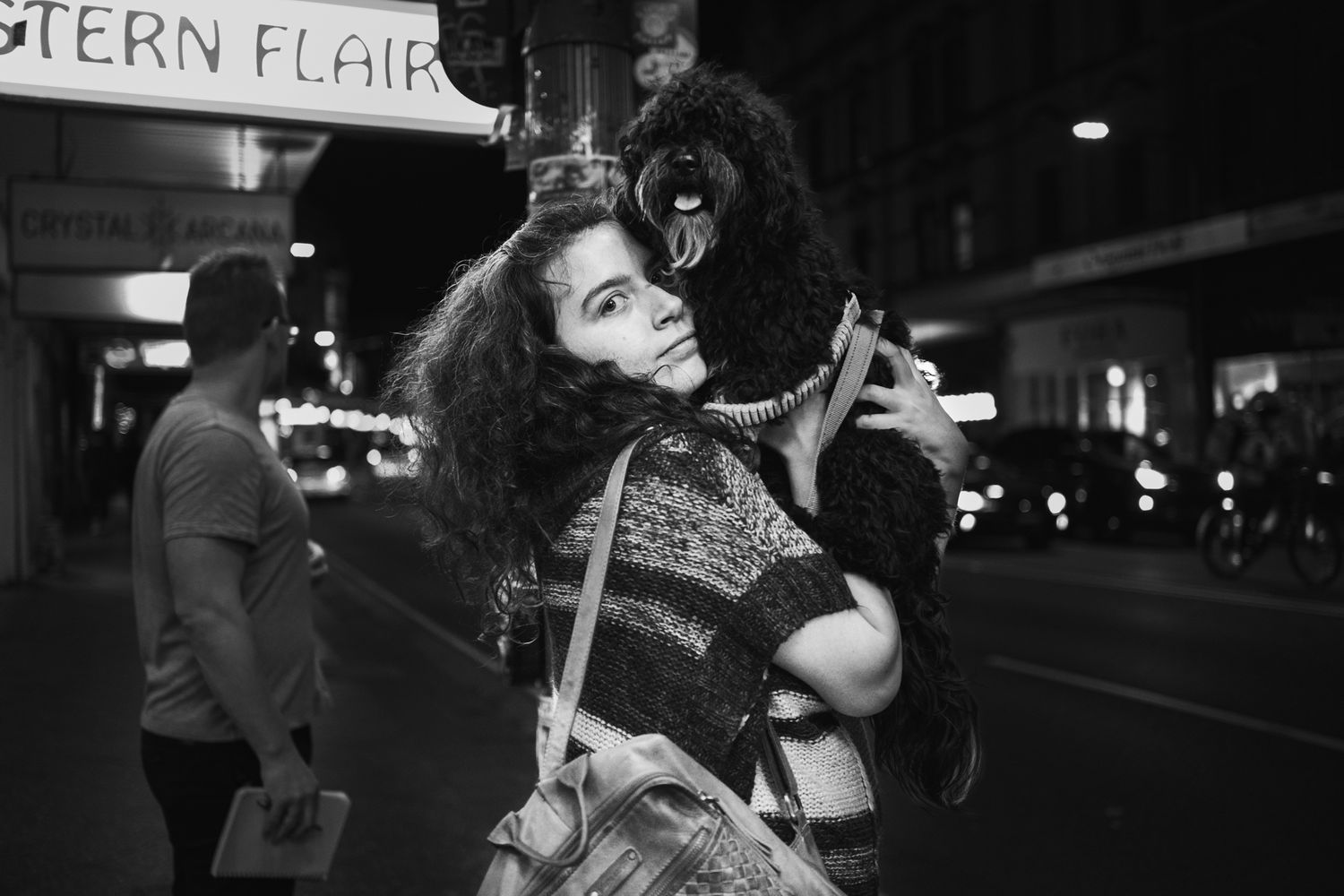 Young woman holds black poodle dog on busy city street at night in black and white photography.