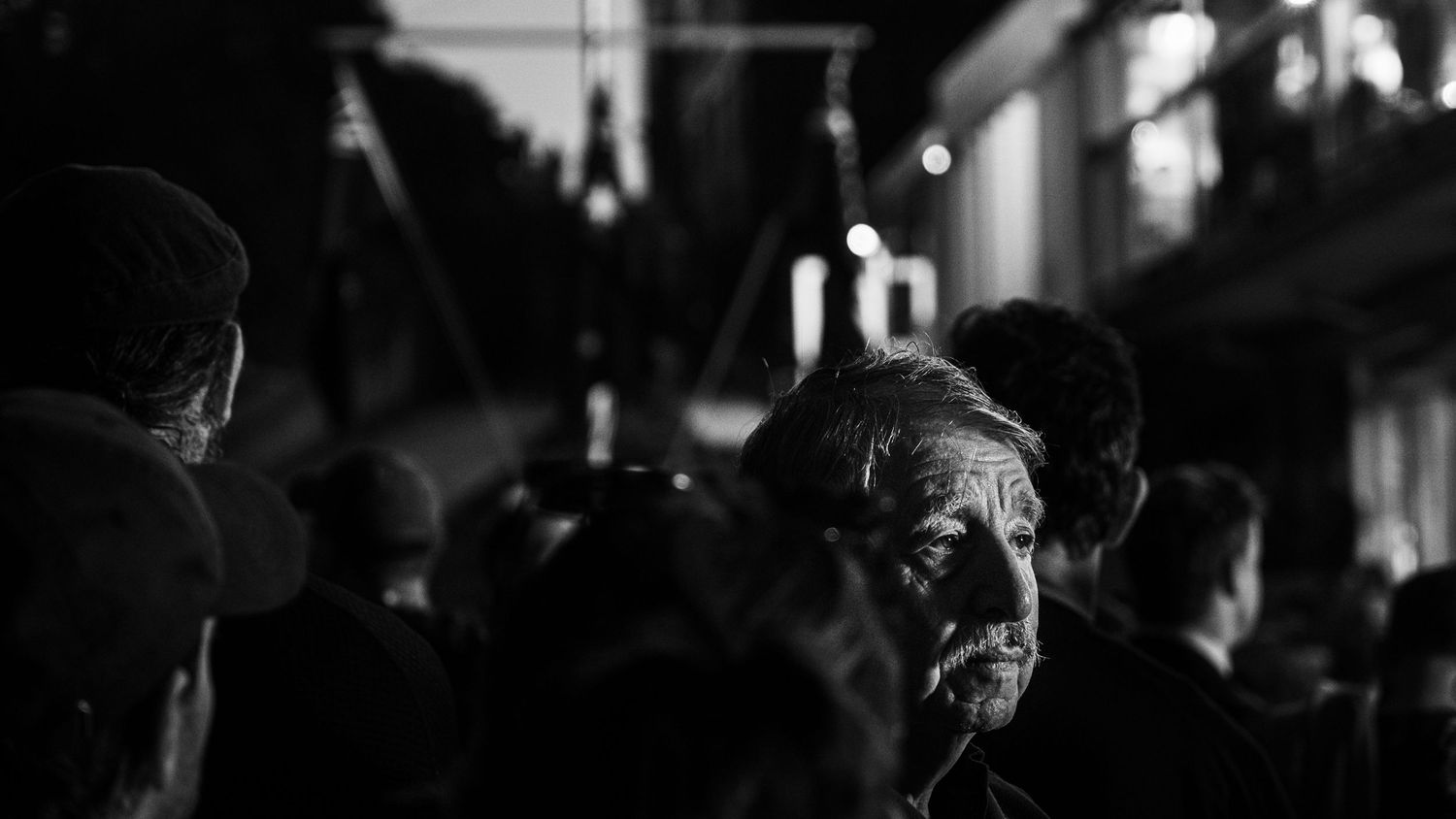 Elderly man with weathered face stands in a crowd at night in a black and white street photography shot.