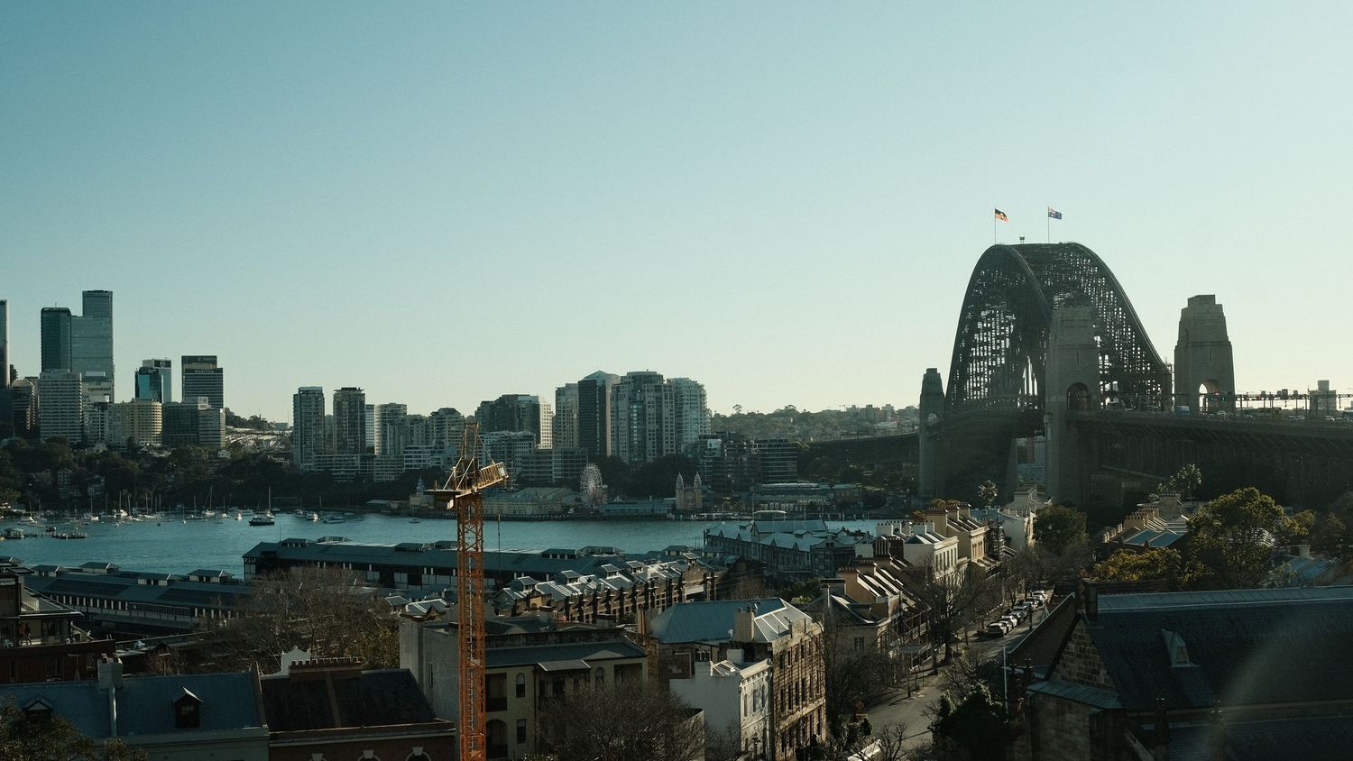 Aerial view of Sydney Harbour Bridge and city skyline on a clear day, with blue waters and construction crane visible.