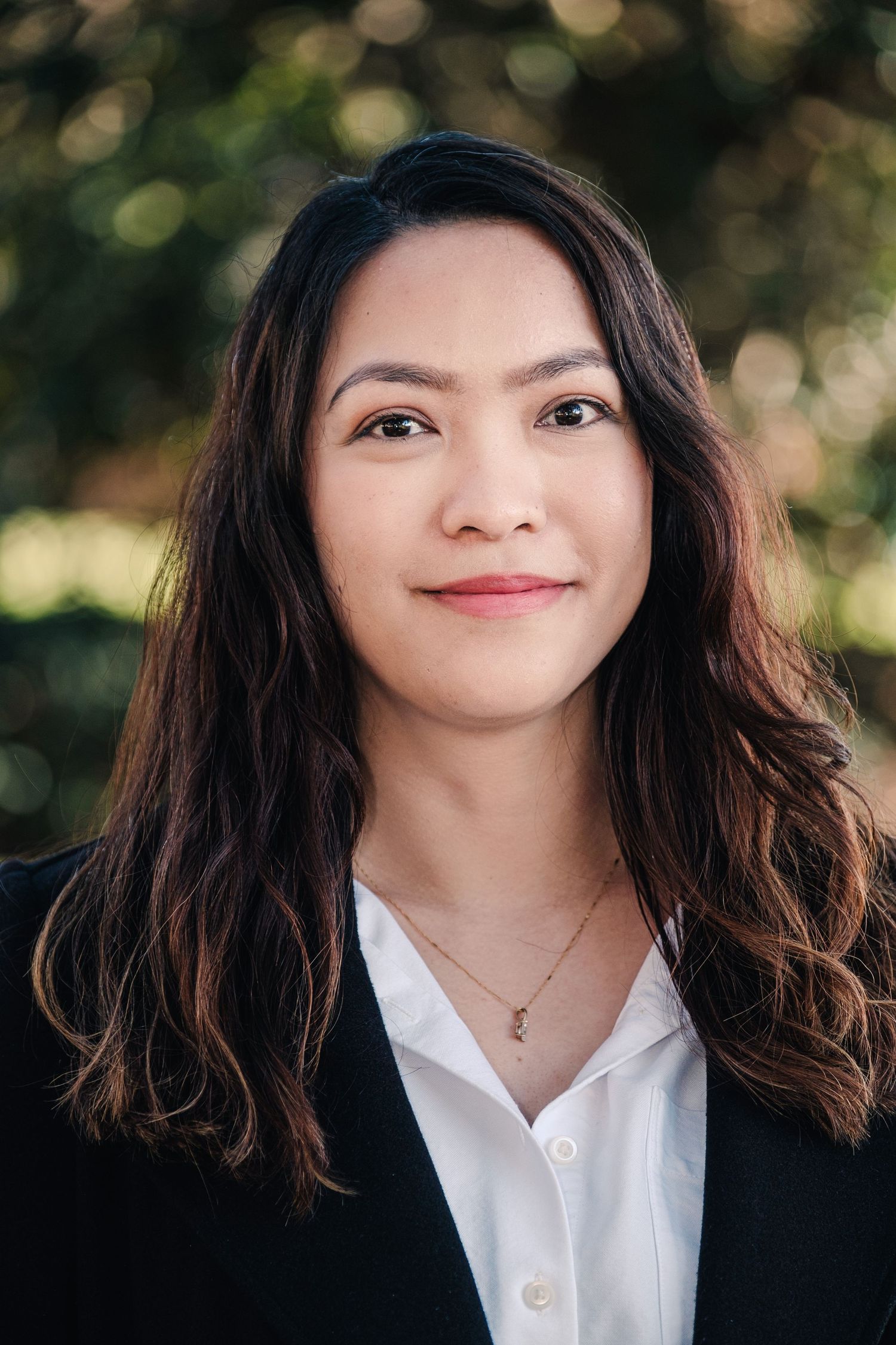 Young woman with long dark hair smiling outdoors, wearing a black blazer and white shirt with a gold necklace.