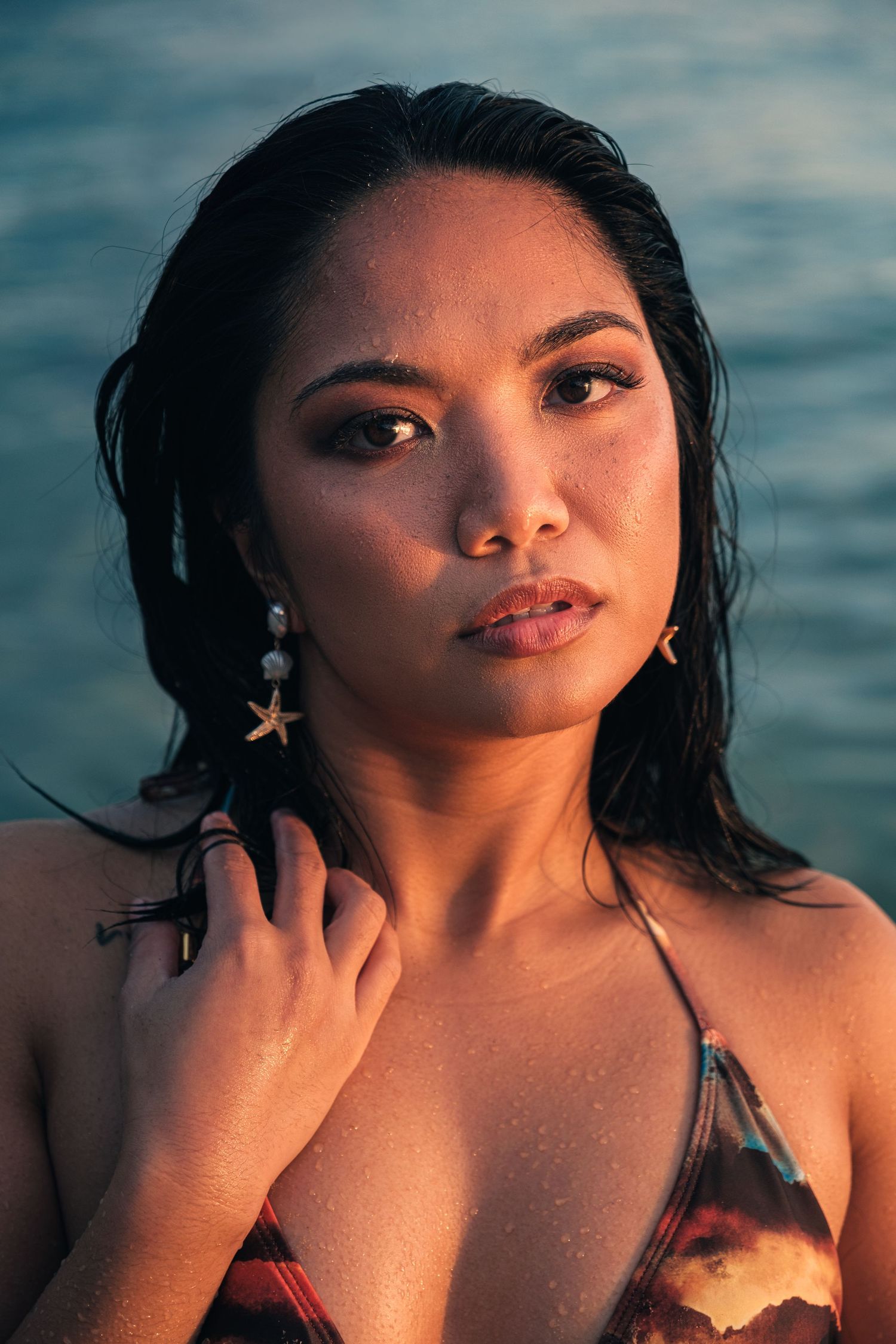 A close-up portrait of a woman wearing starfish earrings with wet hair against a ocean backdrop.