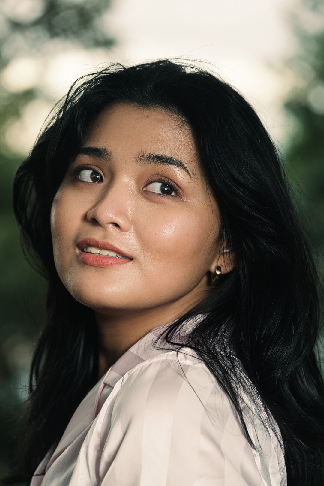 A young Asian woman with long black hair smiles softly outdoors in natural light.