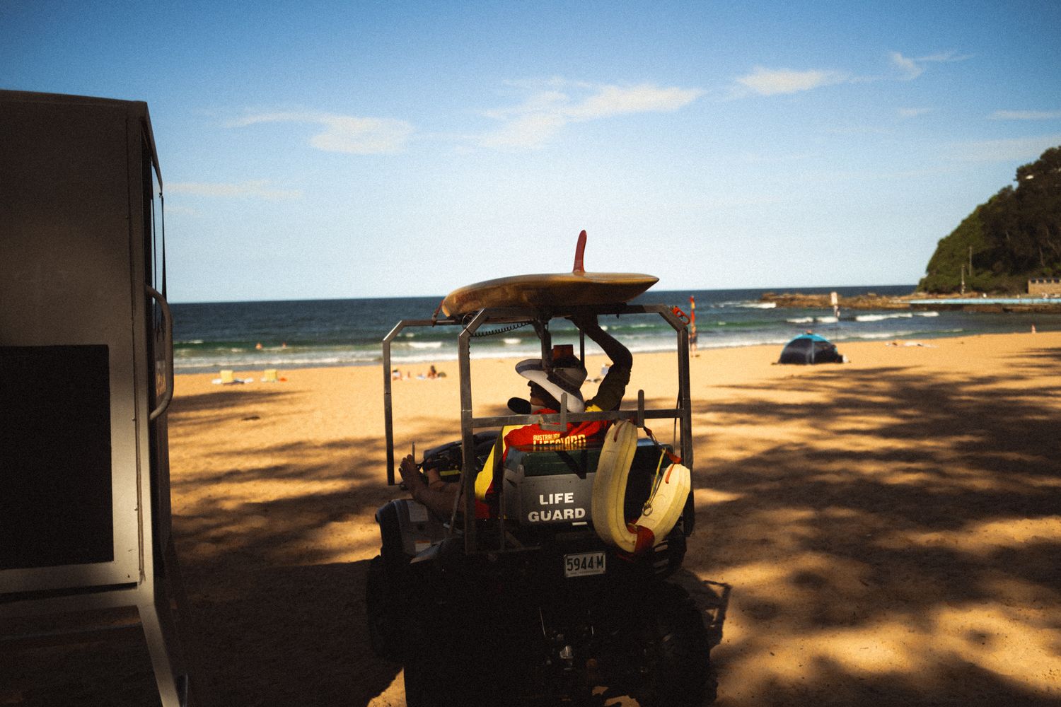 Lifeguard sits on yellow vehicle with surfboard on sunny sandy beach with calm ocean in background.