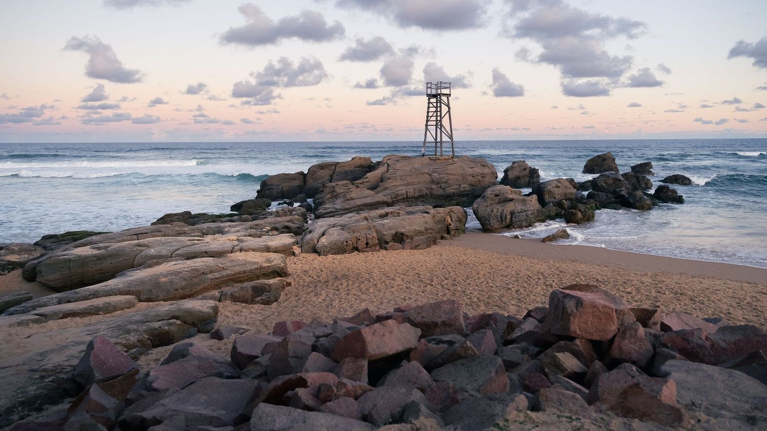 Rocky beach at sunset with a lifeguard tower and gentle ocean waves.
