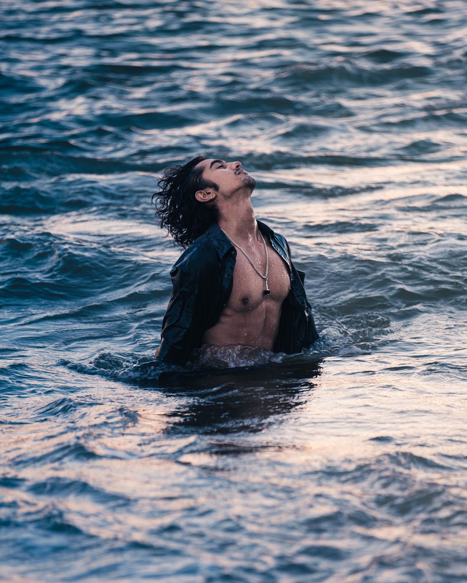 A young man with curly hair emerges from ocean waves wearing an open black jacket.